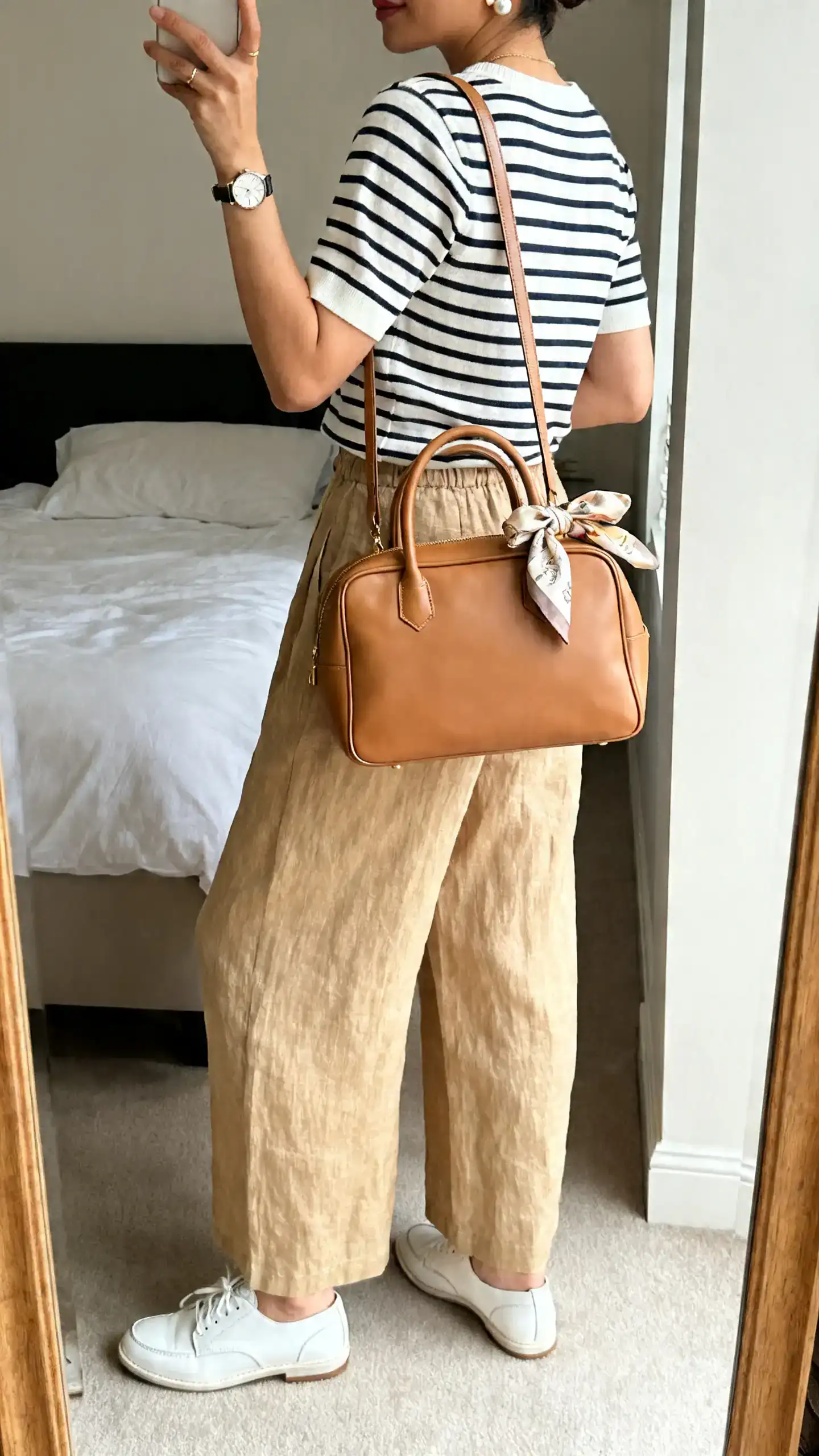 iPhone back-view mirror selfie of a woman wearing wide-leg linen trousers in sand, a Breton striped boat-neck tee, supportive white leather sneakers, a structured tan shoulder bag, classic watch, pearl studs, and a silk scarf tied on the bag handle; face not visible, bedroom mirror, natural light, iPhone photo quality.