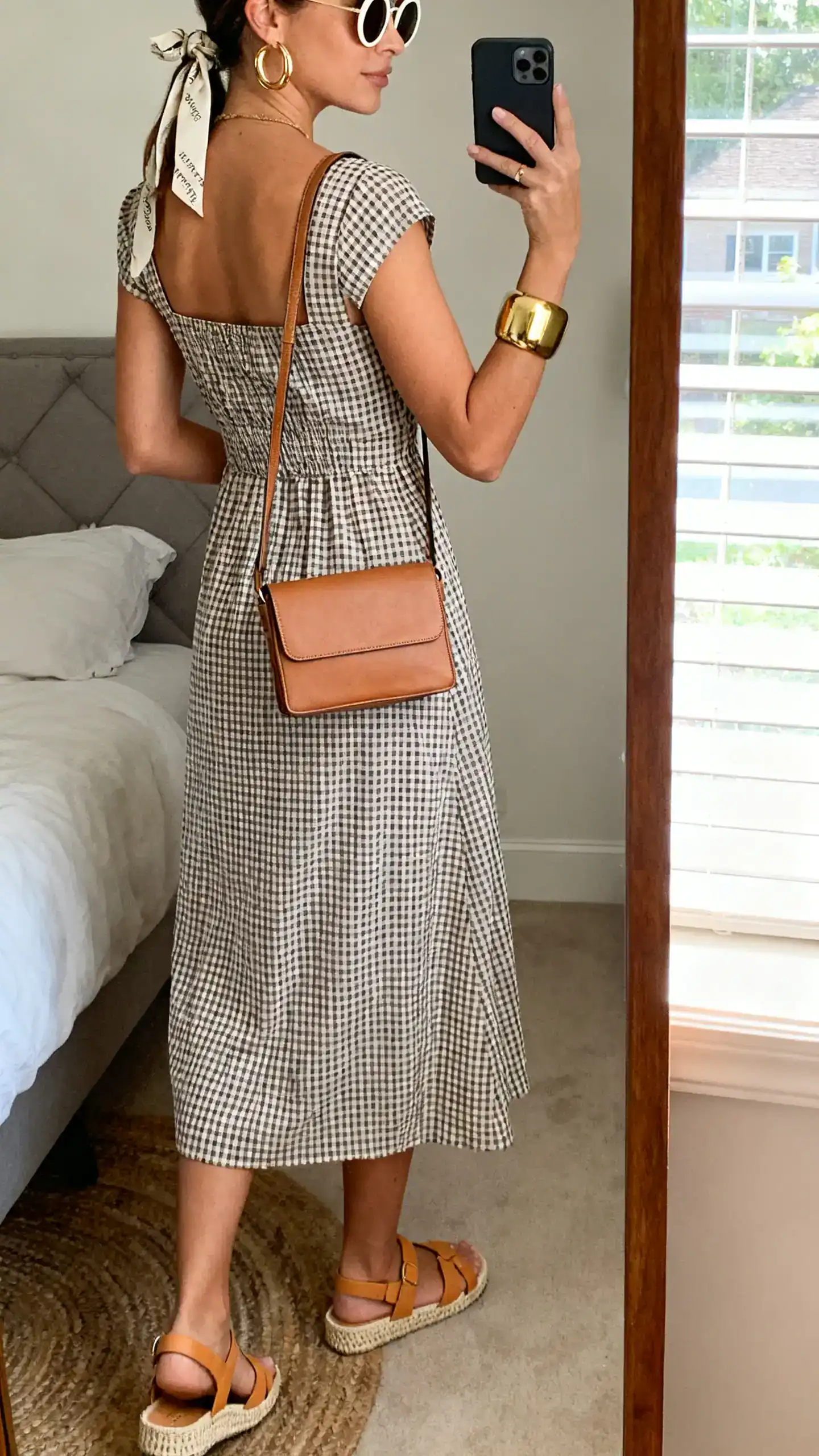 iPhone back-view mirror selfie of a woman wearing a gingham midi sundress with a square neckline, tan fisherman sandals, a leather crossbody with clean lines, chunky gold hoops, a bold cuff bracelet, a hair ribbon, and round sunglasses, face not visible, full-length mirror in bedroom, natural window light, iPhone photo quality.