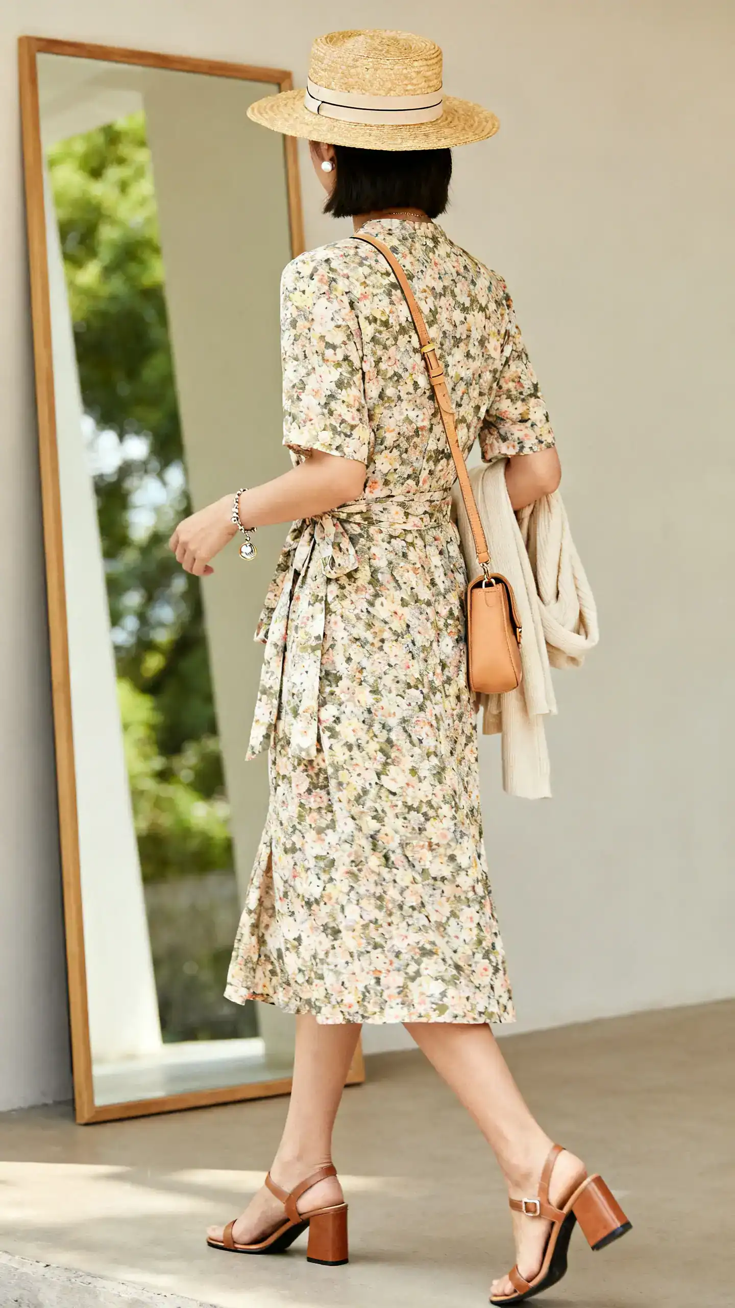 iPhone back view of a woman in a floral wrap midi dress, block-heel sandals, a straw hat with a slim ribbon, a tan leather crossbody, a charm bracelet and classic studs, with a light cardigan over her arm, face not visible, walking pose in front of a full-length mirror, soft afternoon light, iPhone photo quality.