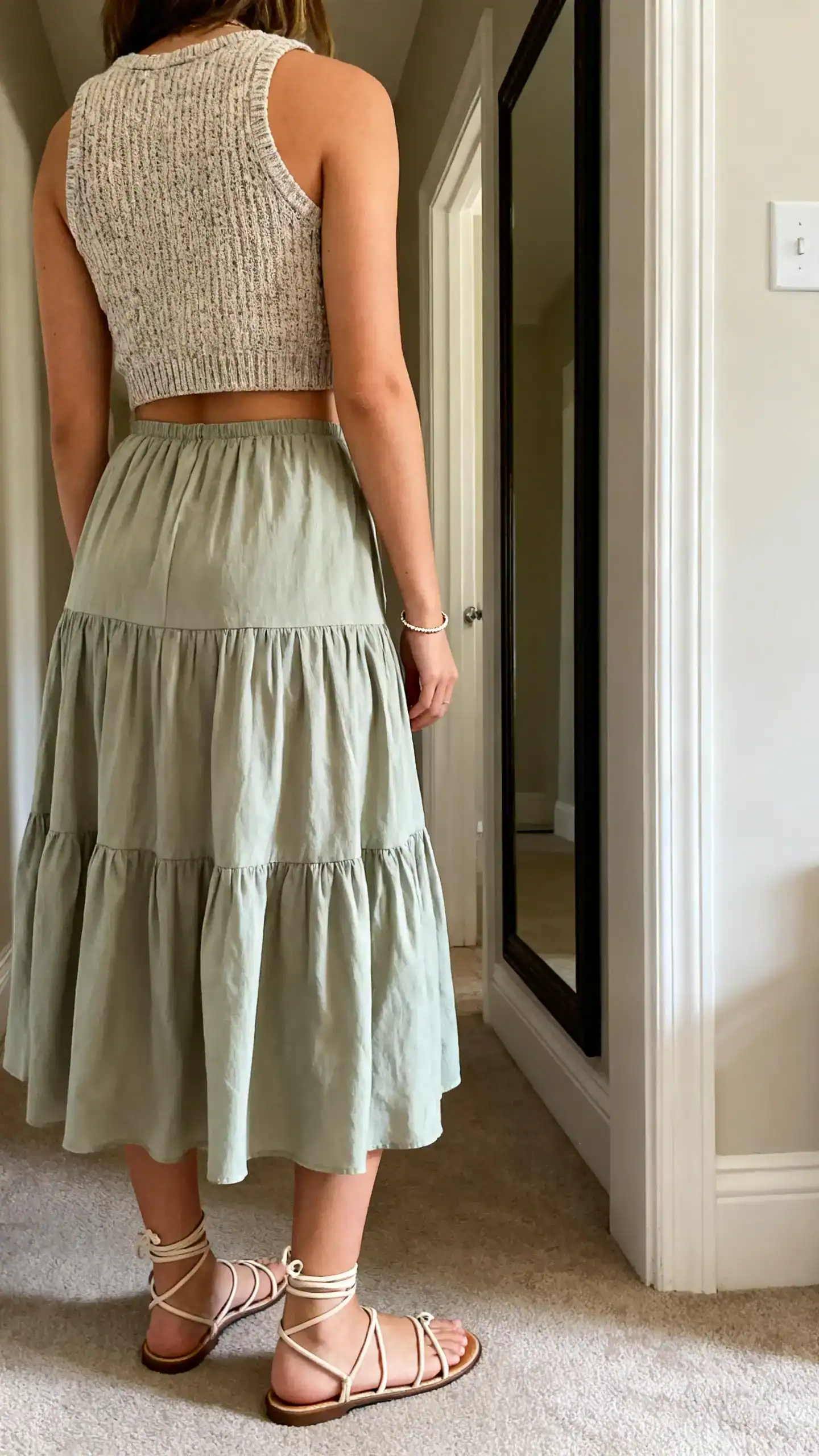 iPhone back-view photo of a woman in a cropped knit tank and a swishy midi skirt with strappy sandals and a delicate bracelet, face not visible, hallway full-length mirror, afternoon indoor light, iPhone photo quality.