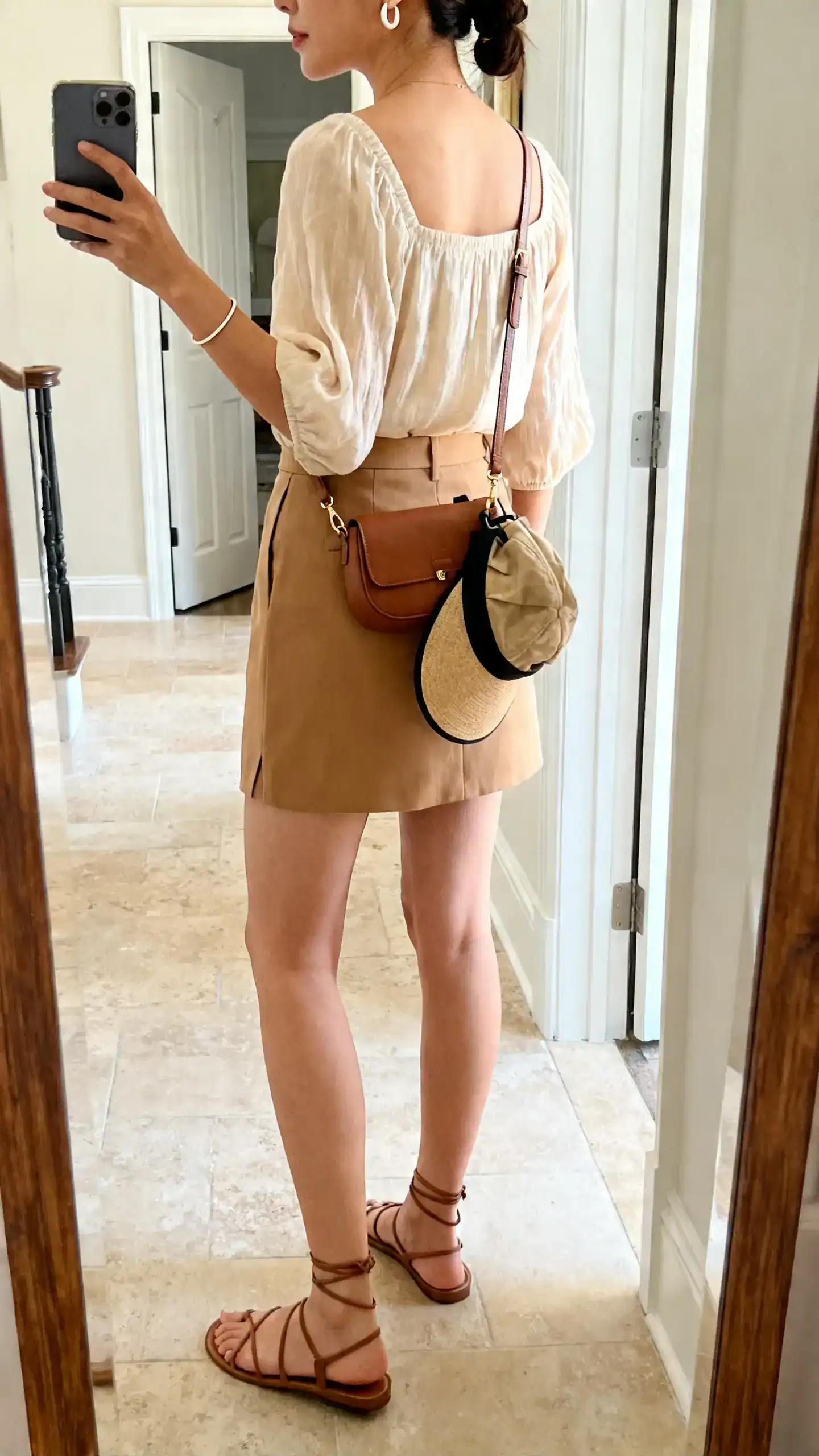 iPhone back-view selfie of a woman in a tailored camel skort, a breezy square-neck blouse, strappy low sandals, a hands-free crossbody, light ear cuffs with a slim bangle, and a packable sun hat clipped to the bag; face not visible, full-length mirror in entryway, natural daylight, iPhone photo quality.
