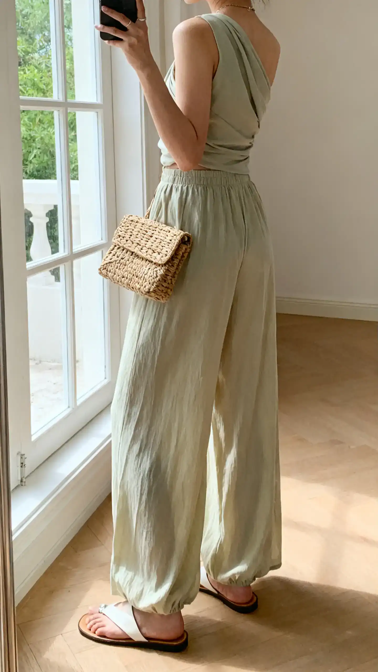iPhone back-view selfie of a woman in breezy palazzo pants with a one-shoulder top, slide sandals, and a woven clutch, face not visible, near window, airy indoor daylight, iPhone photo quality