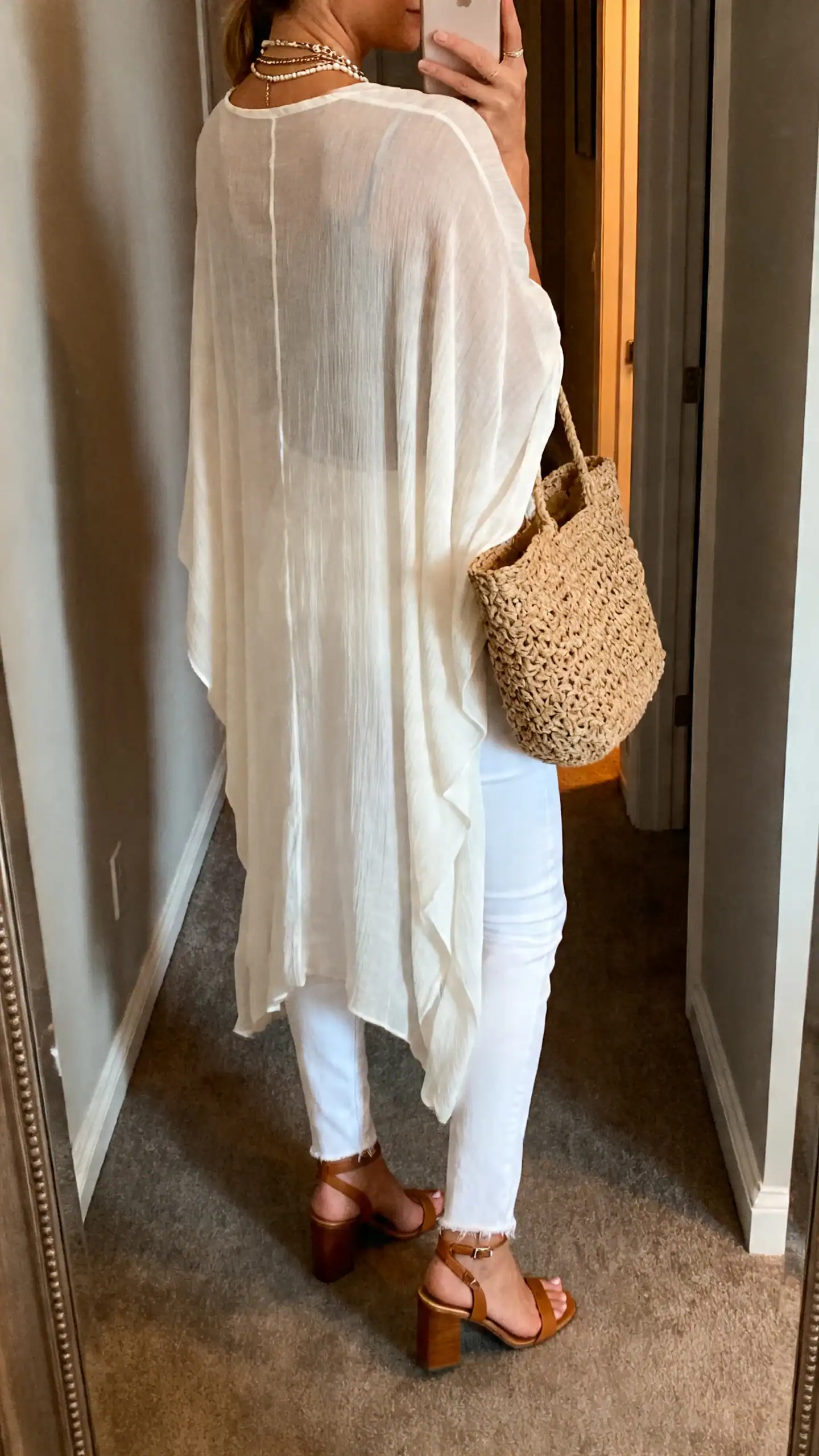 iPhone back-view selfie of a woman in white jeans with a flowing kimono duster, heeled sandals, layered necklaces, woven tote, face not visible, hallway mirror, soft evening light, iPhone photo quality