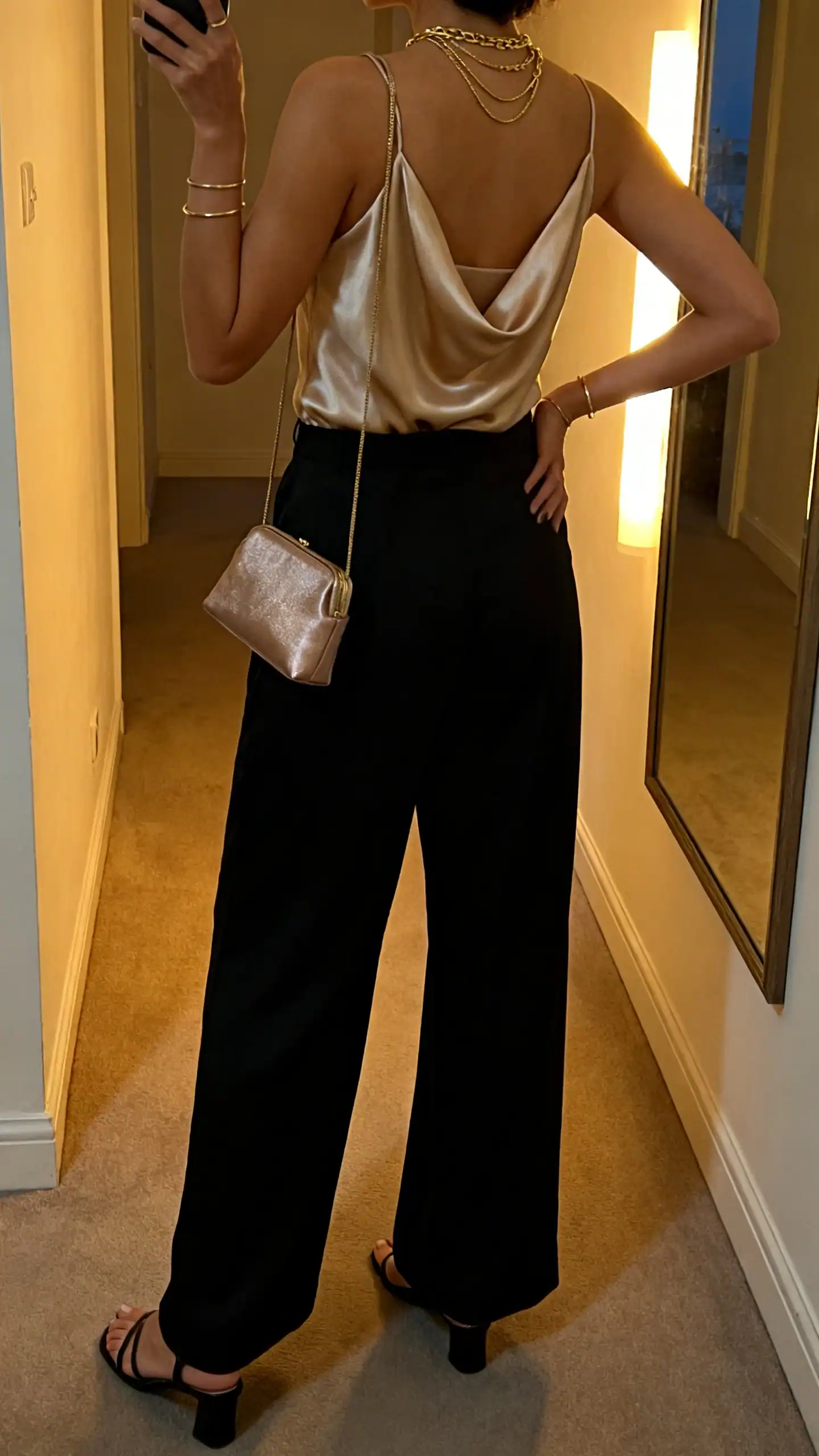 iPhone back-view selfie of a woman wearing a champagne satin-feel cowl-neck cami, black fluid wide-leg trousers, minimal heeled sandals, a mini shoulder bag with subtle shine, layered gold necklaces, and slim bangles; face not visible, hallway mirror at dusk indoor lighting, iPhone photo quality.