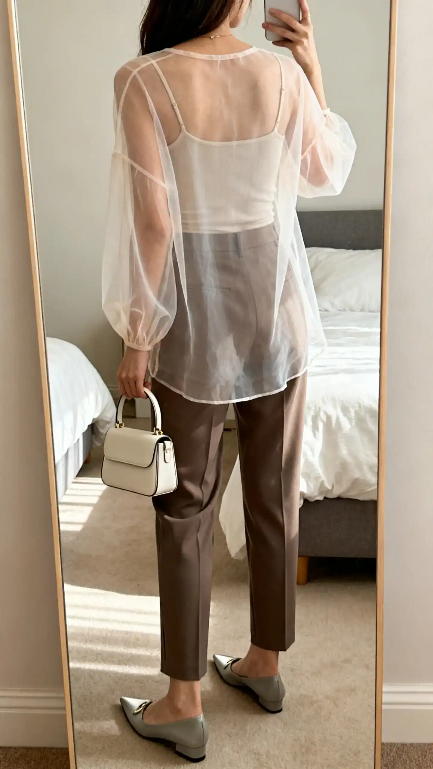 iPhone back-view selfie of a woman wearing a sheer organza top over a fitted camisole, tailored cigarette pants, pointed flats, and a mini top-handle bag, face not visible, full-length mirror in a softly lit bedroom, iPhone photo quality.