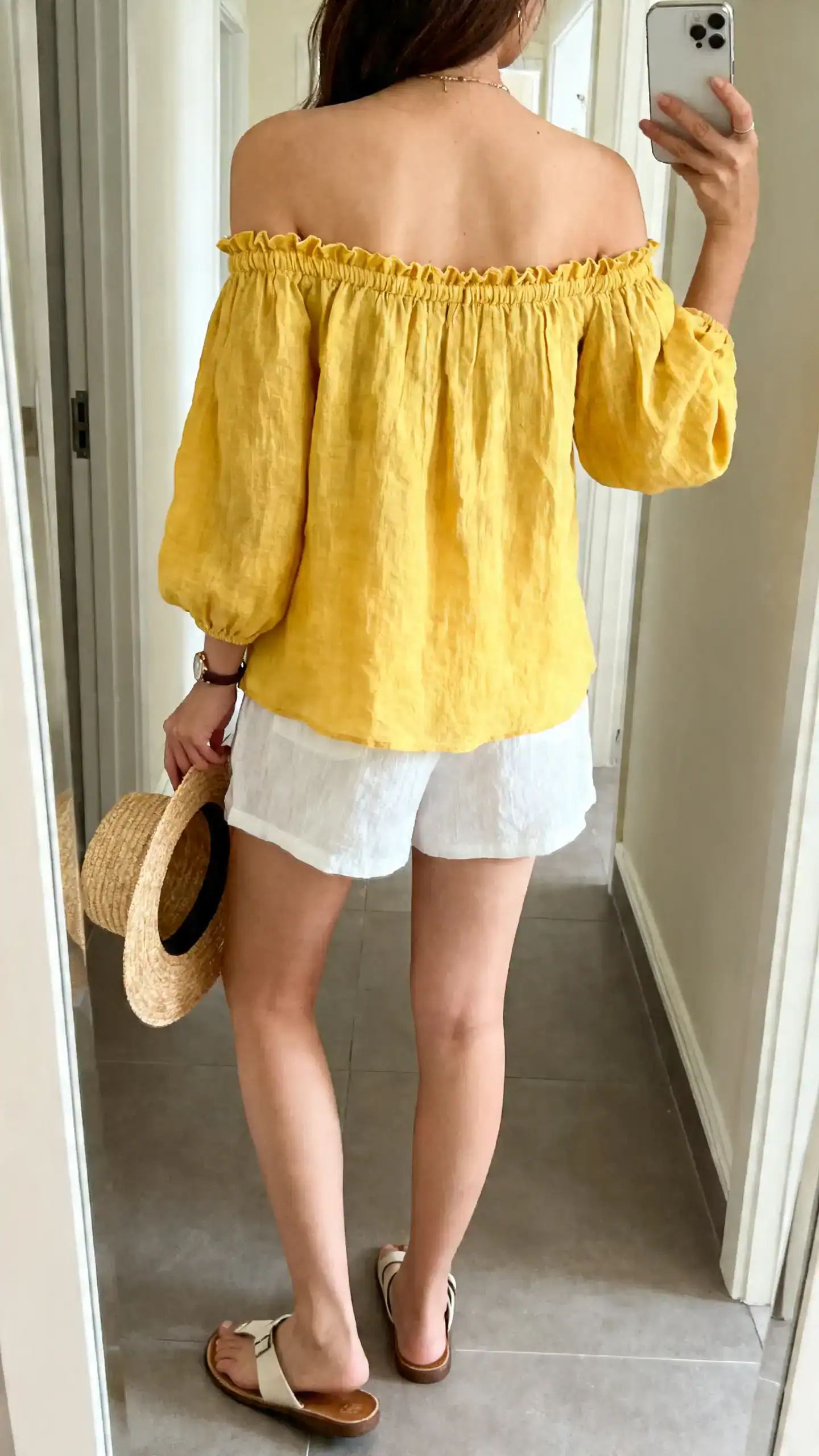 iPhone back-view selfie of a woman wearing a sunny yellow off-shoulder linen top with delicate elastic neckline, white linen shorts, flat sandals, straw hat in hand, face not visible, hallway mirror, bright natural light, iPhone photo quality