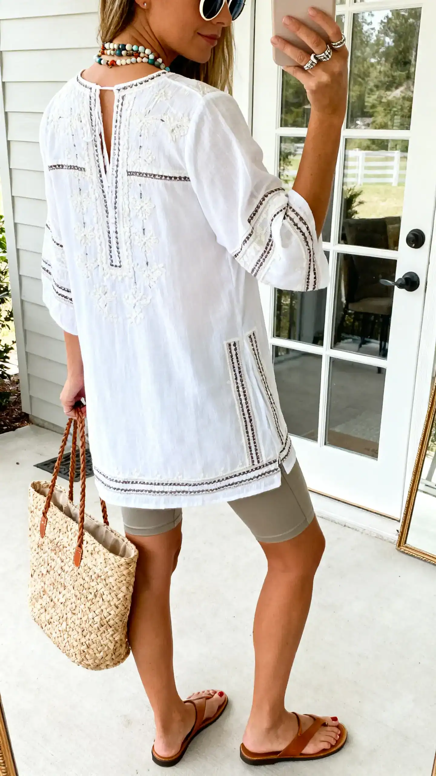 iPhone back-view selfie of a woman wearing a white embroidered tunic with contrast detailing, slim-fit cutoff shorts, toe-post leather sandals, woven carryall tote, layered beaded necklaces, stackable rings, and round-frame sunglasses; face not visible, patio door mirror, bright daylight, casual iPhone photo.