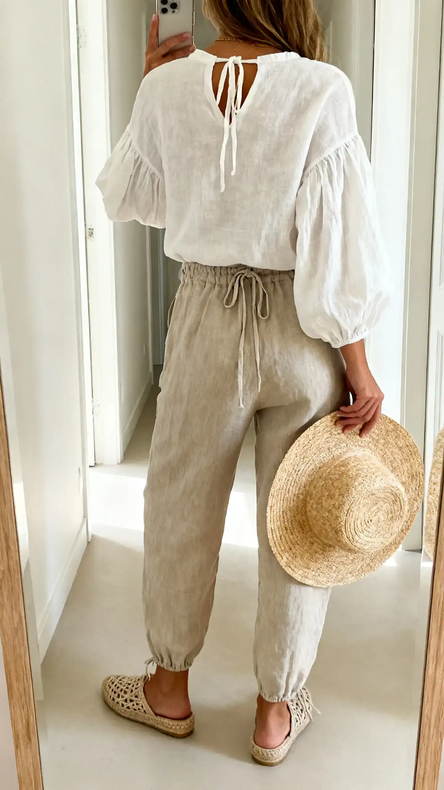 iPhone back-view selfie of a woman wearing an off-white linen poet blouse with gathered sleeves and tie neckline, relaxed linen drawstring trousers, woven espadrille flats, and a raffia sunhat in hand, face not visible, full-length mirror in a bright hallway with natural daylight, iPhone photo quality.