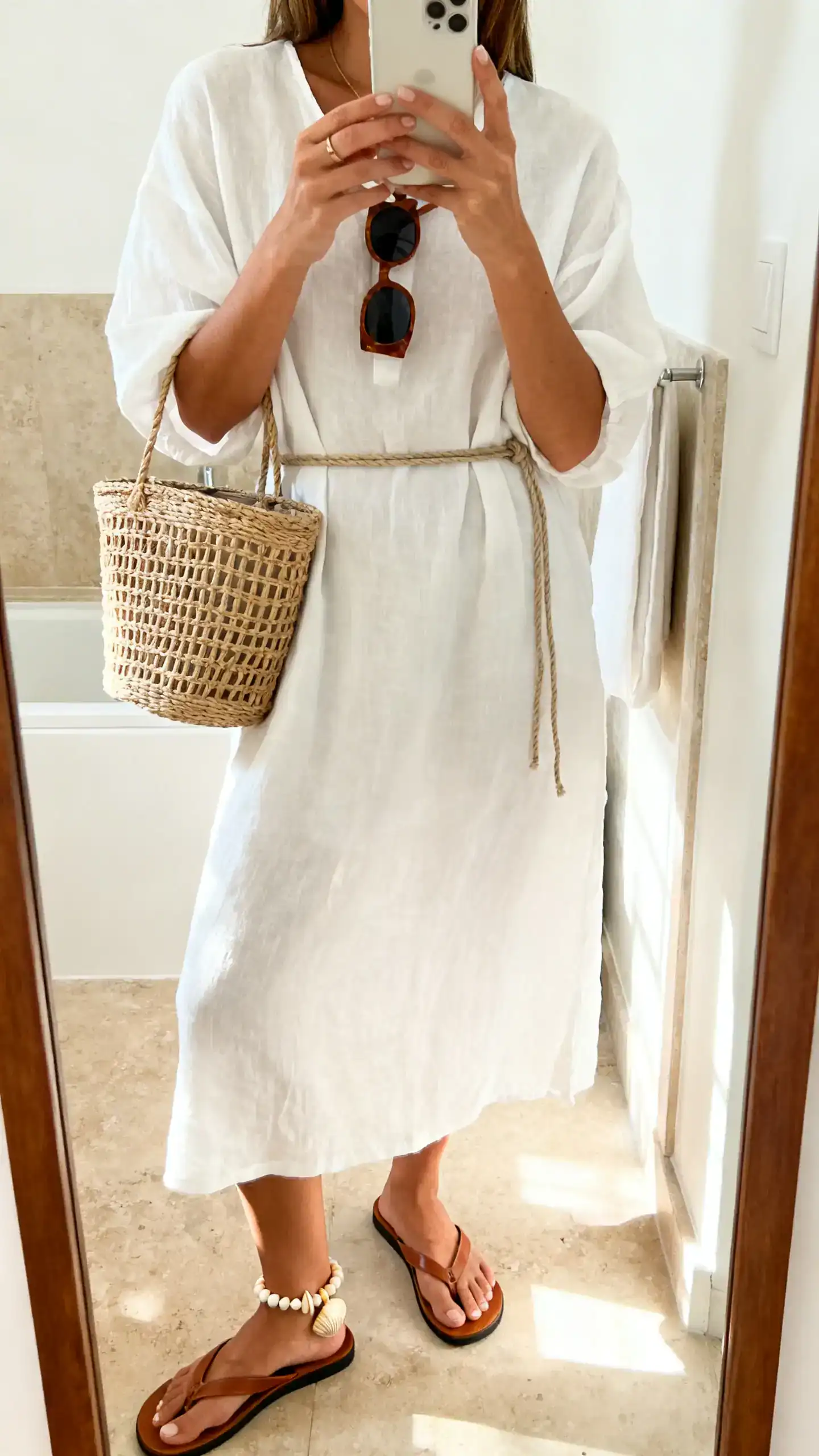iPhone casual mirror selfie of a woman in a slightly oversized white cotton gauze dress (midi) with leather flip-flops, woven bucket bag, shell/beaded anklet, oversized sunglasses, optional thin rope belt; face not visible, bathroom mirror, bright morning light, iPhone photo quality.