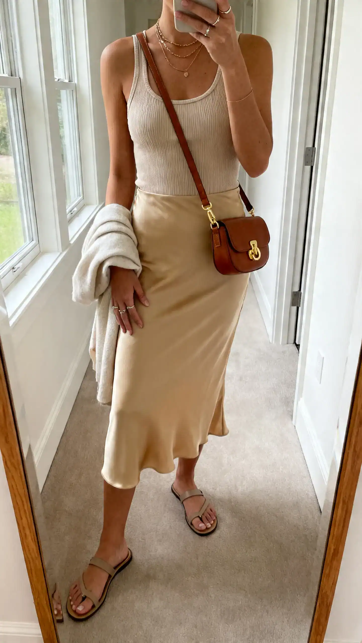 iPhone mirror selfie of a woman in a bias-cut midi slip skirt in champagne with a fitted ribbed tank, sleek flat sandals, leather crossbody with gold hardware, layered necklaces and delicate rings, and a light wrap over one arm; face not visible, cropped at shoulders, hallway full-length mirror, soft window light, iPhone photo quality.