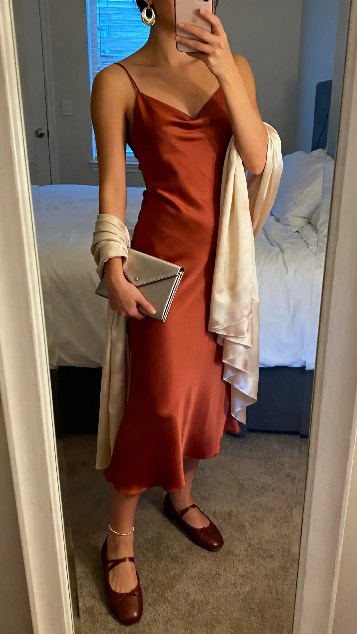 iPhone mirror selfie of a woman in a bias-cut slip dress in terracotta, leather ballet flats, a slim envelope micro clutch, refined statement earrings, a light cashmere-silk wrap over one arm, and a subtle anklet, face not visible, cropped at neck, bedroom mirror, soft evening light, iPhone photo quality.