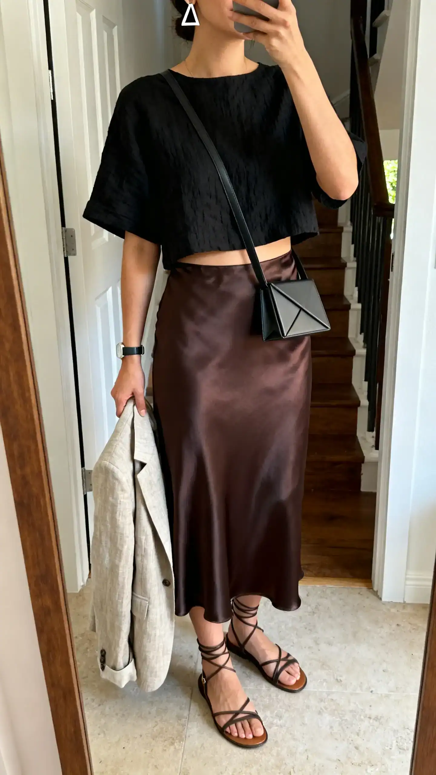 iPhone mirror selfie of a woman in a black boxy cropped linen-blend top, high-waisted satin midi slip skirt in complementary tone, strappy flat sandals, an angular crossbody bag, geometric earrings, minimalist watch, and a lightweight linen blazer draped over arm; face not visible, cropped at neck, entryway mirror, natural afternoon light, iPhone photo quality.