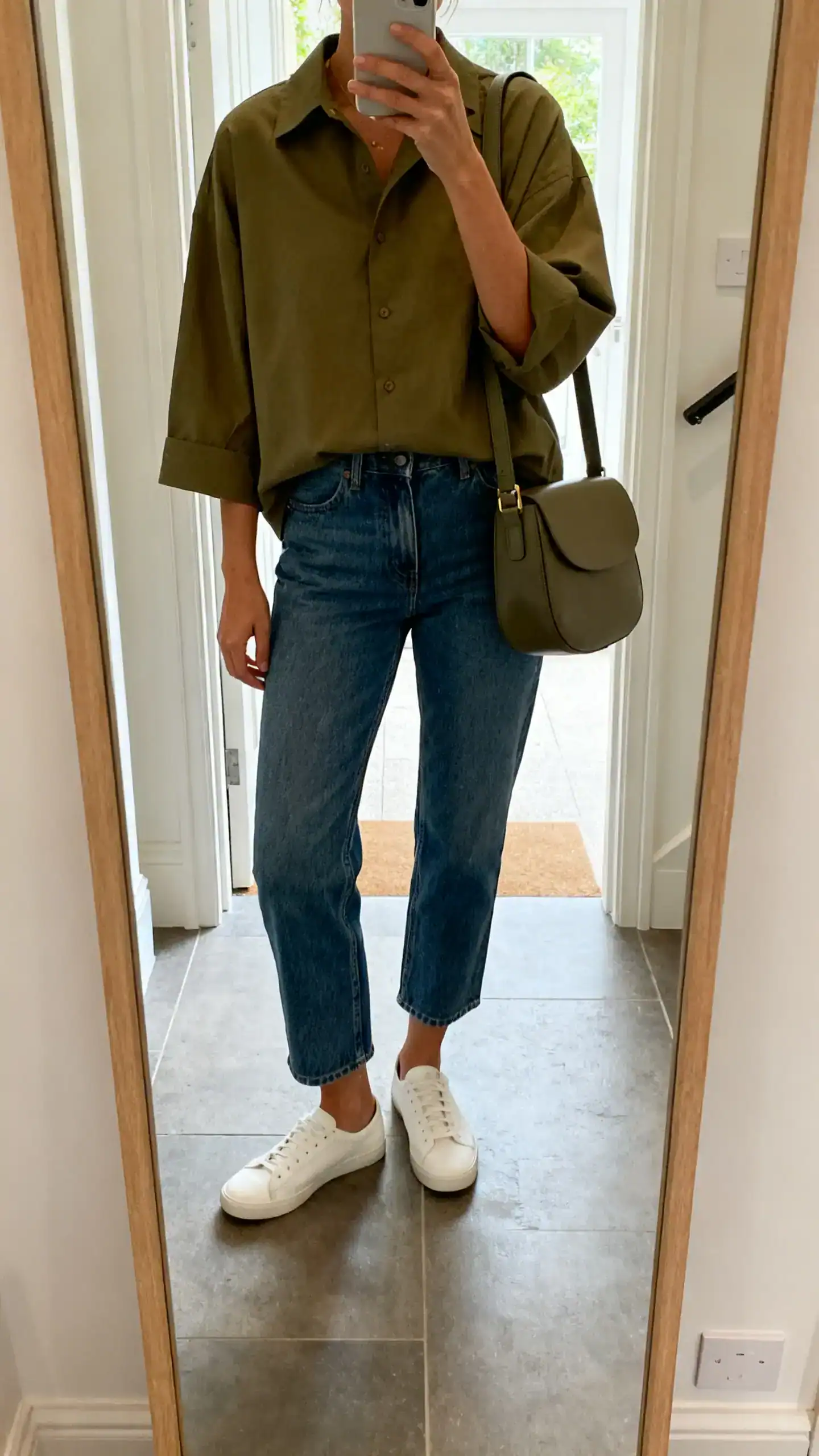 iPhone mirror selfie of a woman in a boxy camp-collar shirt in olive, straight-leg cropped jeans, minimalist white sneakers, and a structured shoulder bag, face not visible, cropped at shoulders, entryway mirror with natural daylight, iPhone photo quality.
