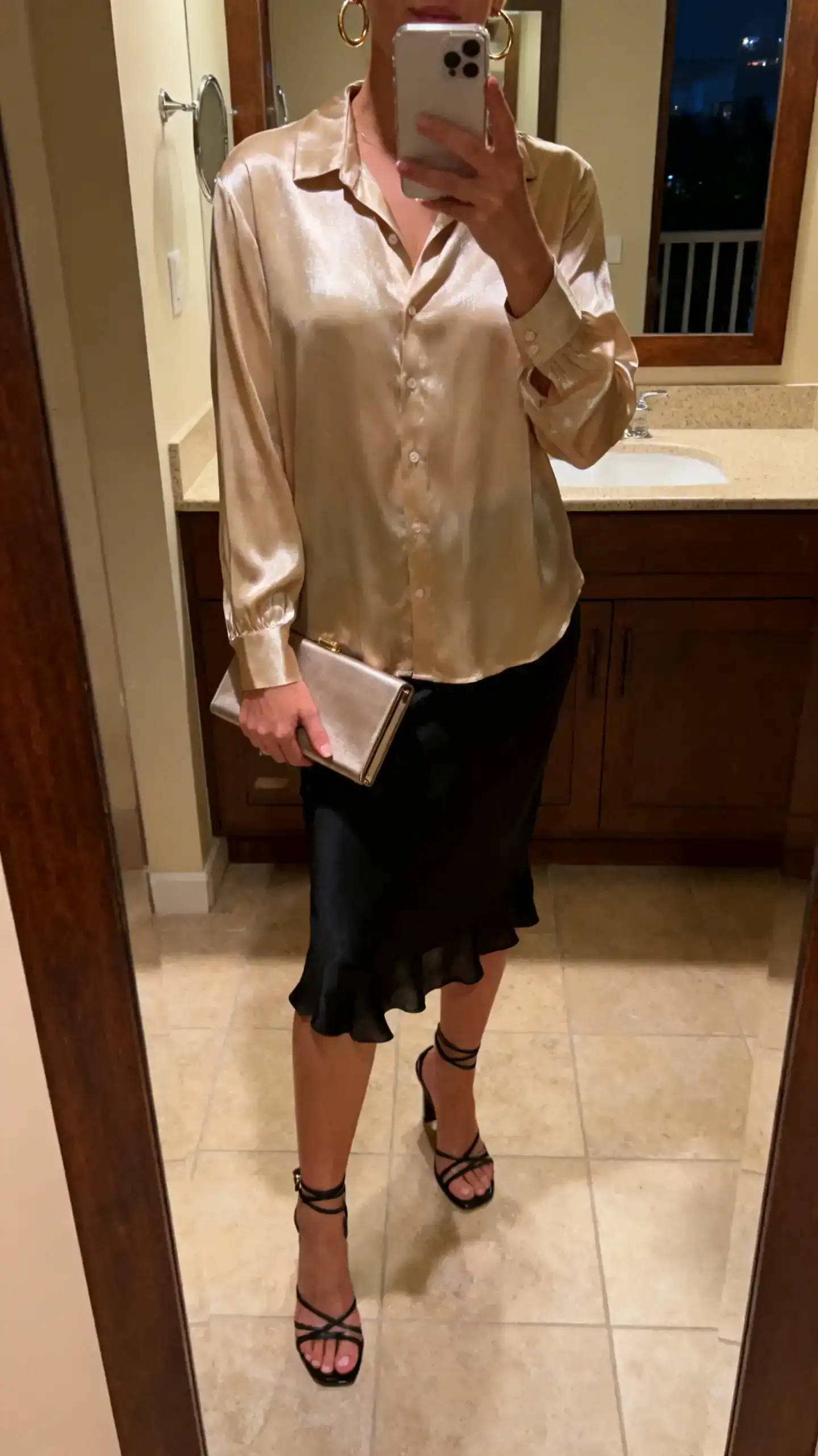 iPhone mirror selfie of a woman in a champagne lightweight satin blouse with subtle sheen, black slip skirt, strappy heeled sandals, a slim clutch, and hoop earrings, face not visible, cropped at neck, bathroom mirror with warm evening indoor lighting, iPhone photo quality.