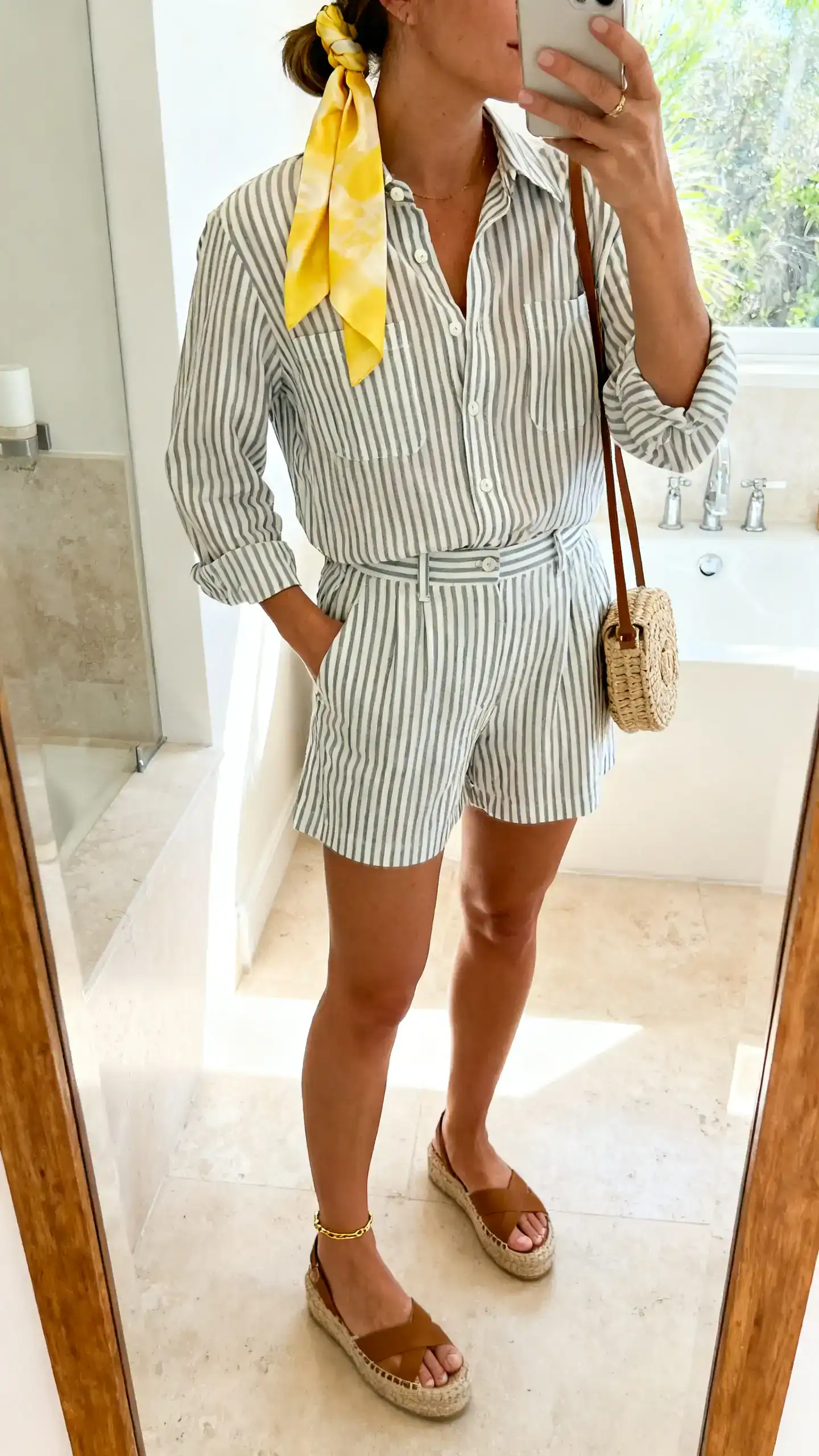 iPhone mirror selfie of a woman in a coordinated striped button-up shirt with matching striped shorts, espadrille wedges, a silk scarf in lemon tied in her hair, small woven shoulder bag, and a subtle gold anklet, face not visible, side angle crop at shoulders, bathroom mirror, bright morning light, iPhone photo quality.