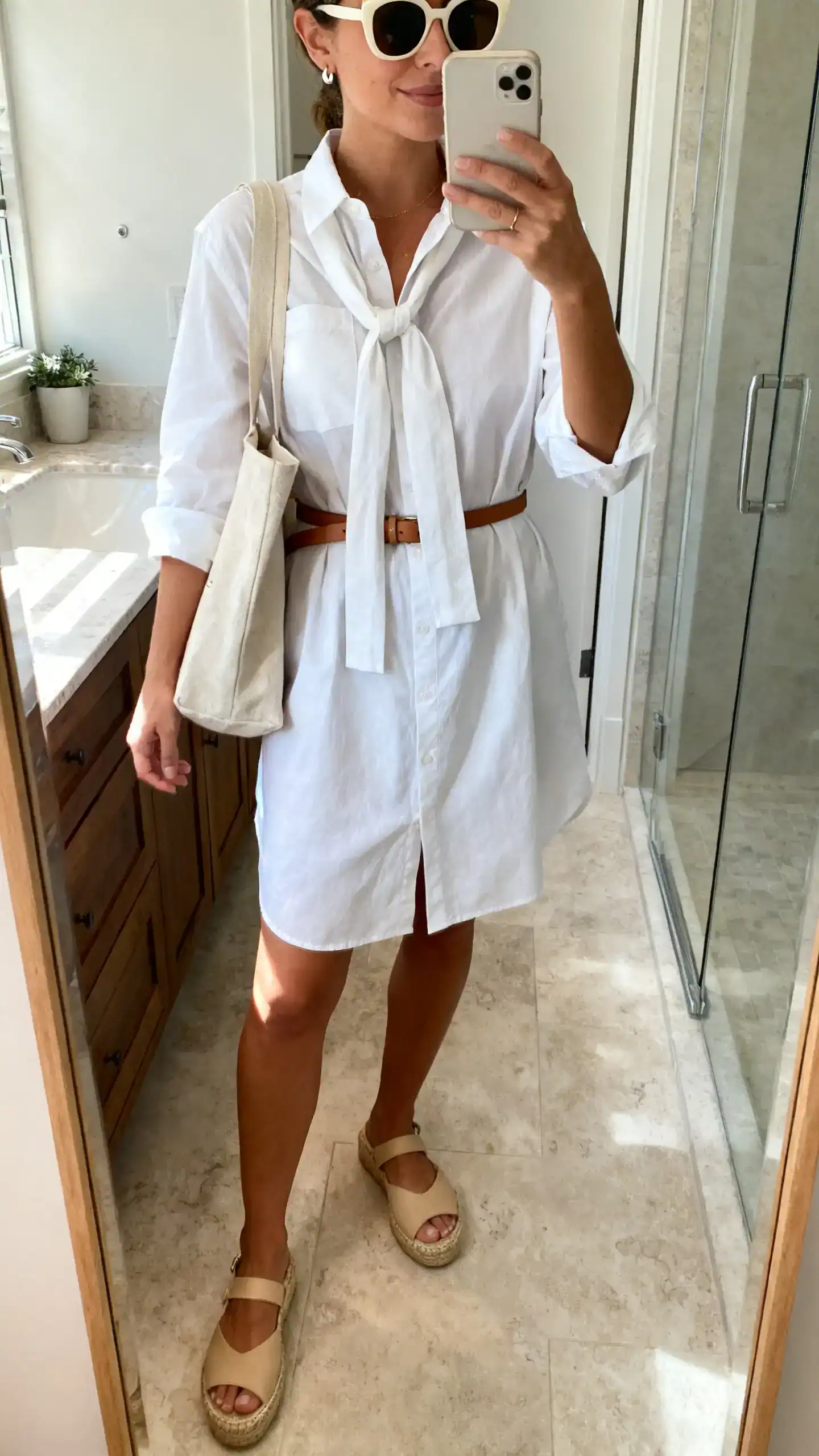 iPhone mirror selfie of a woman in a cotton poplin shirt dress with a removable tie, espadrille wedges in sand, a canvas tote, oversized sunglasses, a thin leather belt, and simple stud earrings, face not visible, bathroom mirror, indoor daylight, iPhone photo quality.