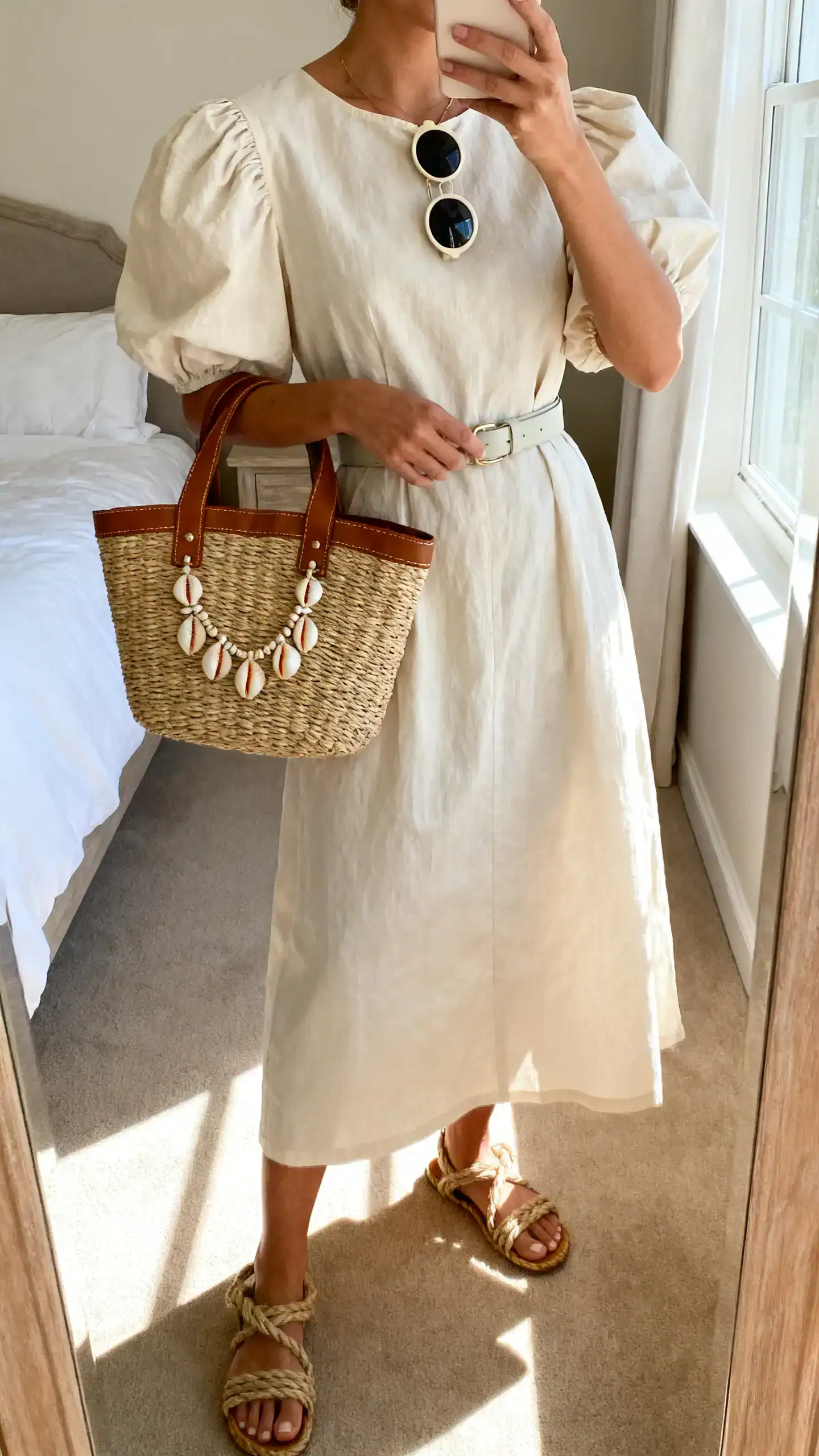iPhone mirror selfie of a woman in a cotton poplin midi dress with puff sleeves, rope sandals, a basket bag with leather trim, shell jewelry accents, round sunglasses, and a light belt at the waist, face not visible, cropped at neck, bedroom mirror, sunny window light, iPhone photo quality.