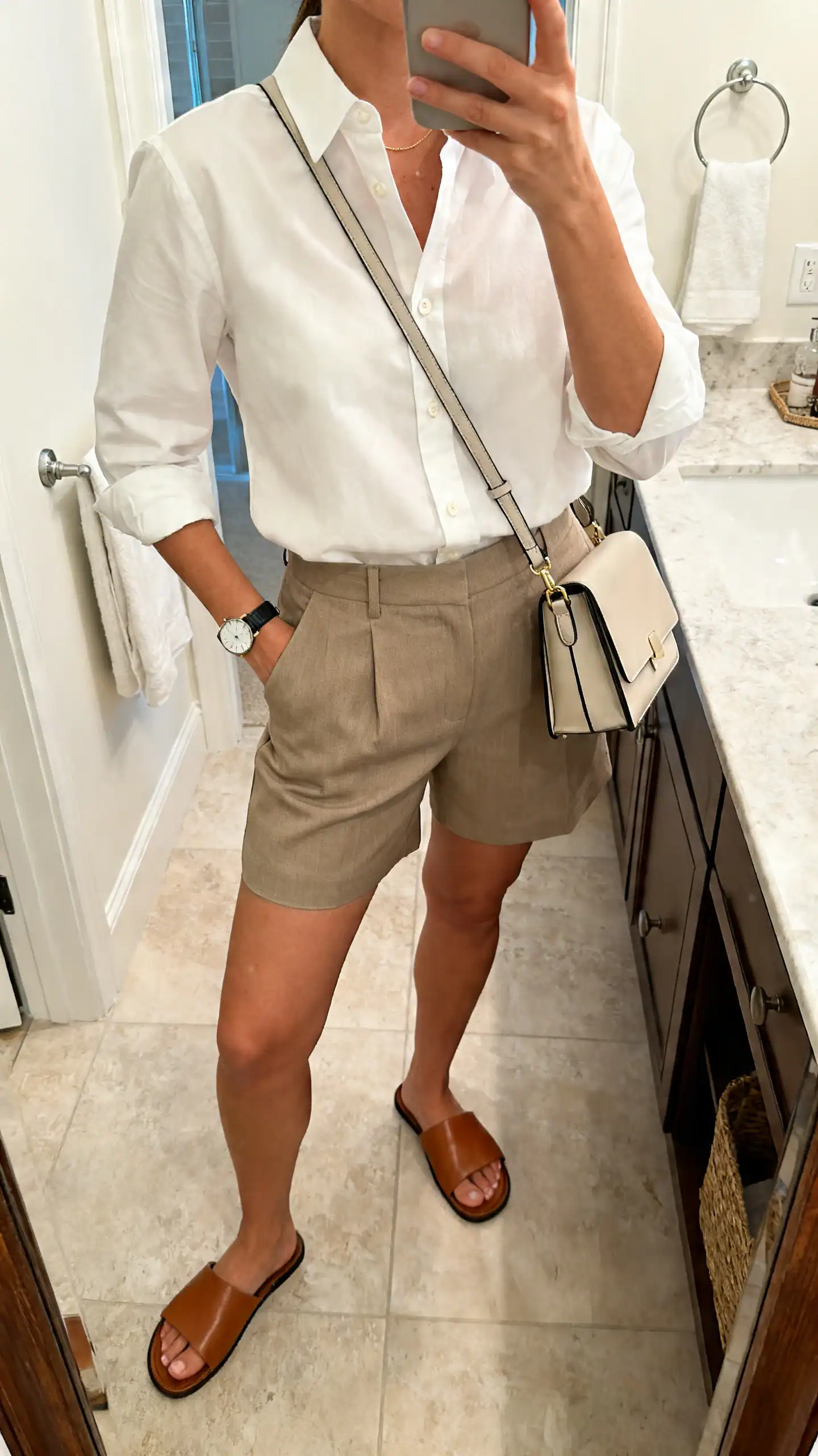 iPhone mirror selfie of a woman in a crisp white button-down tucked into tailored shorts, leather slides, structured crossbody, simple watch, face not visible, bathroom mirror, soft indoor light, iPhone photo quality