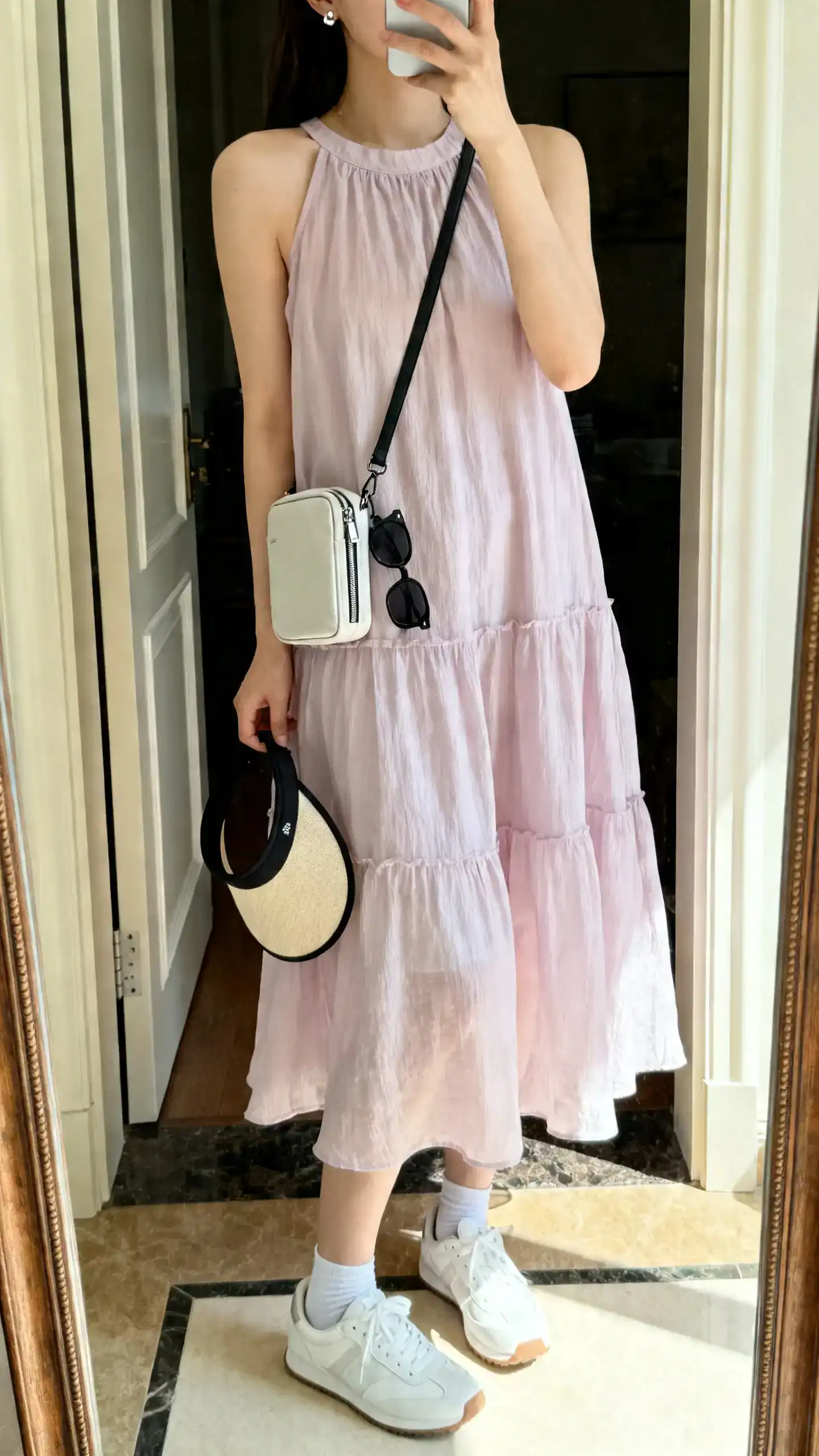 iPhone mirror selfie of a woman in a floaty pastel A-line sundress, low-profile white sneakers, compact zip crossbody, sporty-chic sunglasses, a visor hooked to the bag, minimal stud earrings, and thin no-show ankle socks; face not visible, cropped at neck, entryway mirror, daylight, iPhone photo quality.