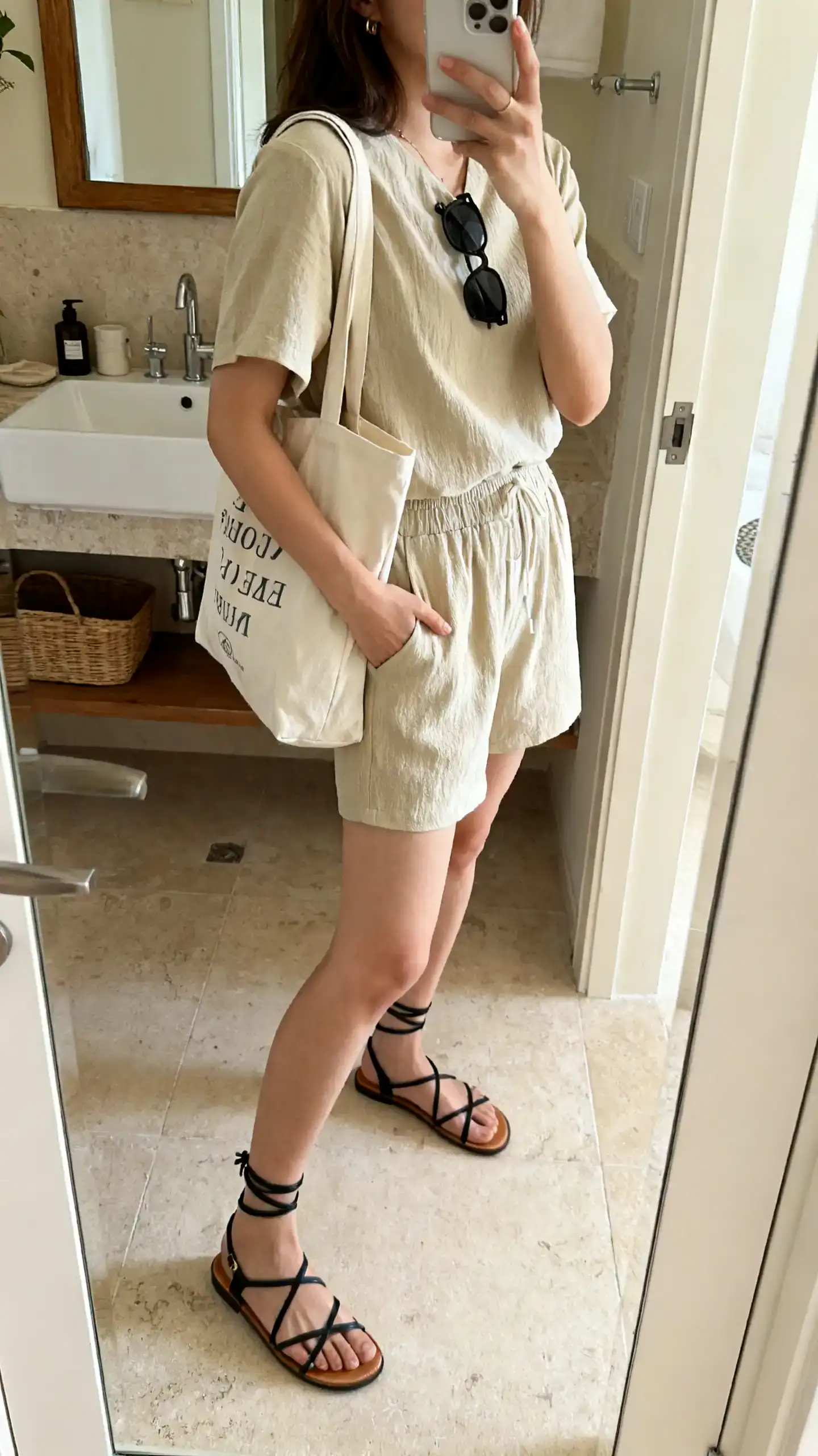 iPhone mirror selfie of a woman in a matching shorts co-ord set with strappy flat sandals, tote bag, and sunglasses, face not visible, side angle, bathroom mirror, soft indoor light, iPhone photo quality