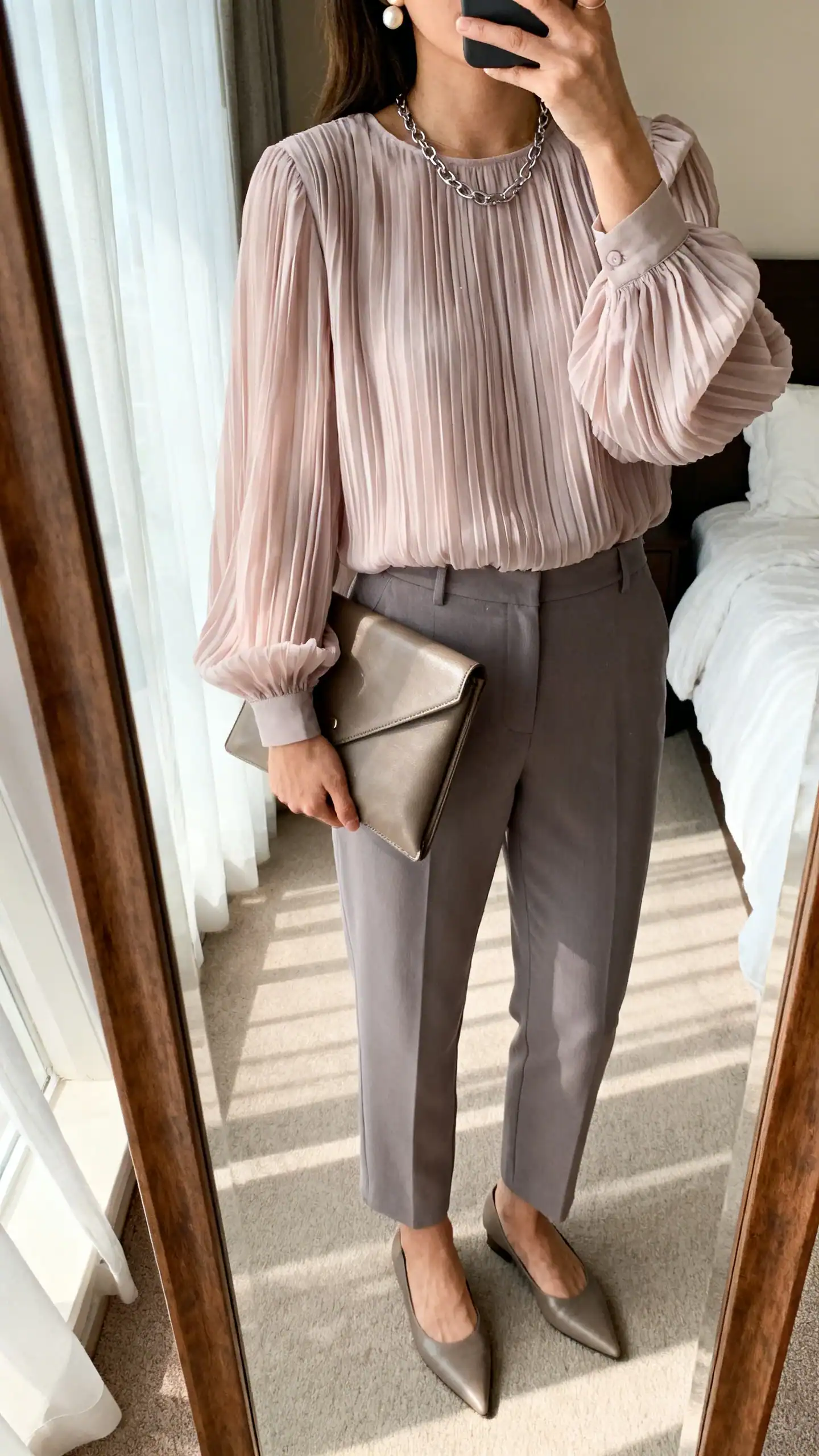 iPhone mirror selfie of a woman in a pastel pleated chiffon blouse with gentle blouson sleeves, tailored ankle-length trousers in dove grey, pointed-toe flats, a leather envelope clutch, pearl studs, and a fine linked chain necklace; face not visible, cropped at shoulders, bedroom mirror, soft window light, iPhone photo quality.
