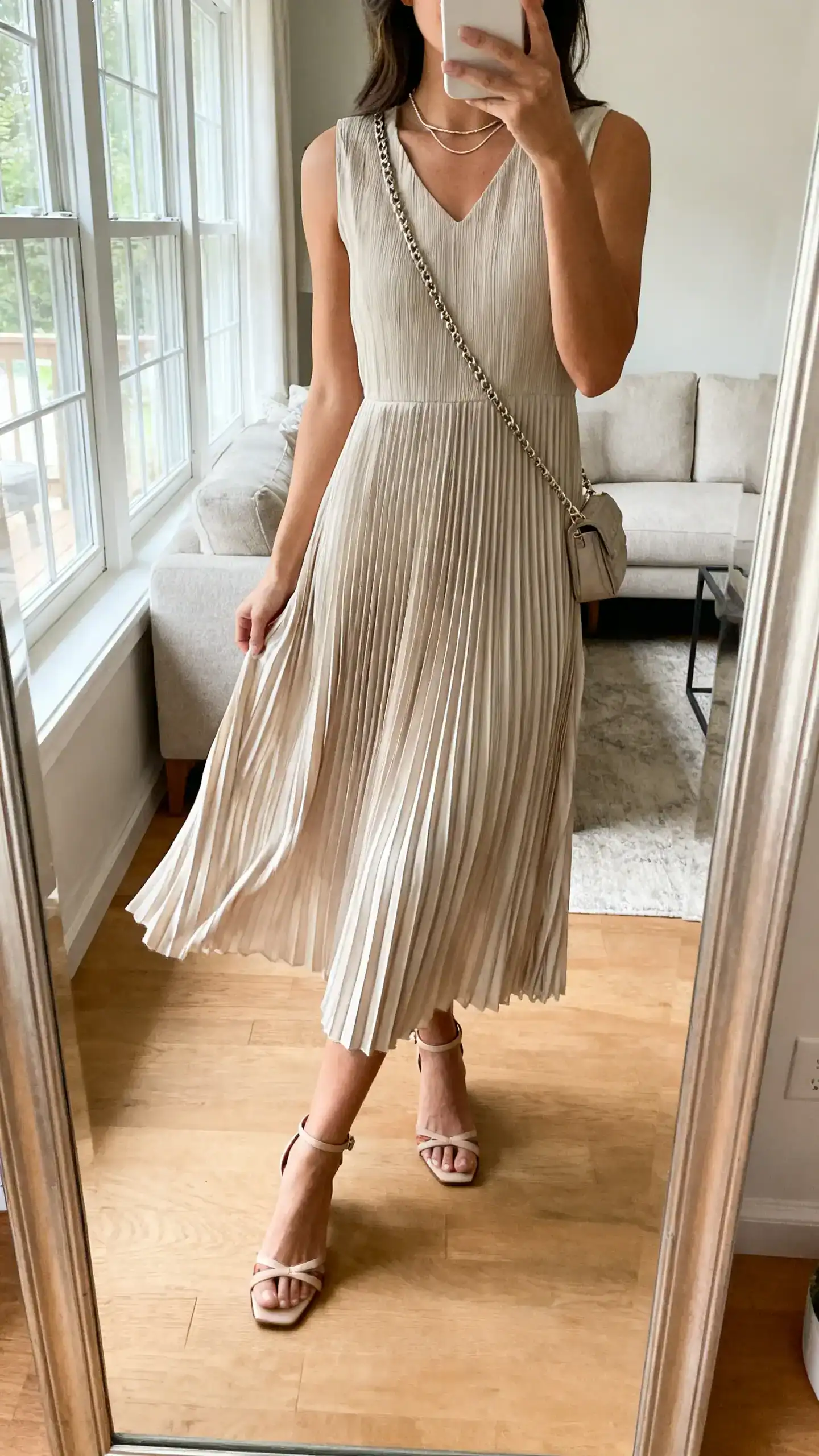 iPhone mirror selfie of a woman in a pleated midi dress that sways, ankle-strap heels, thin chain necklace, and a small shoulder bag, face not visible, cropped at neck, living room full-length mirror, natural window light, iPhone photo quality