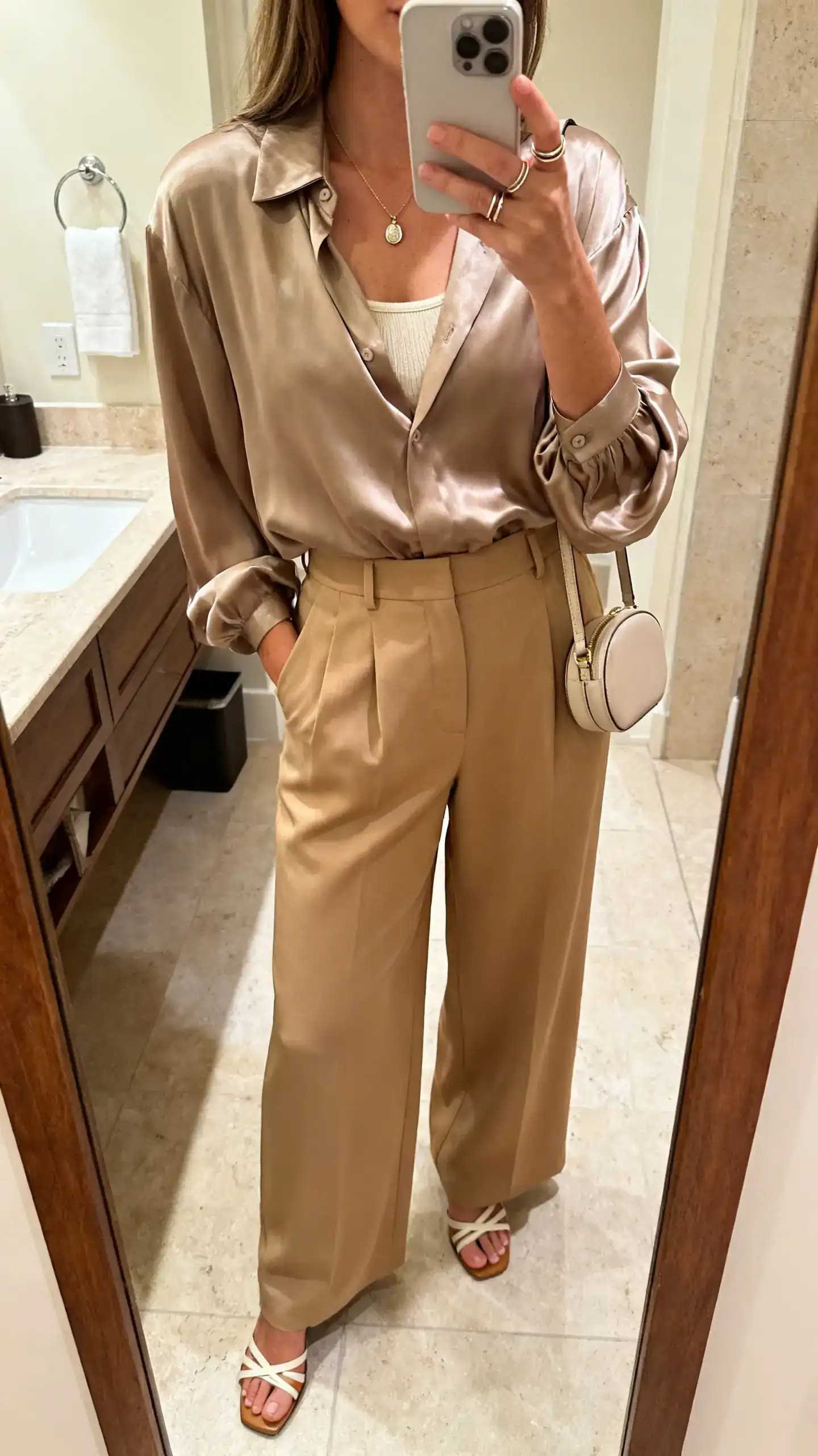 iPhone mirror selfie of a woman in a relaxed satin blouse with soft drape, high-rise wide-leg trousers in sand, strappy low-heel sandals, delicate pendant necklace with thin stacking rings, compact shoulder bag, optional lightweight camisole peeking slightly, face not visible, bathroom mirror, warm indoor lighting, iPhone photo quality.