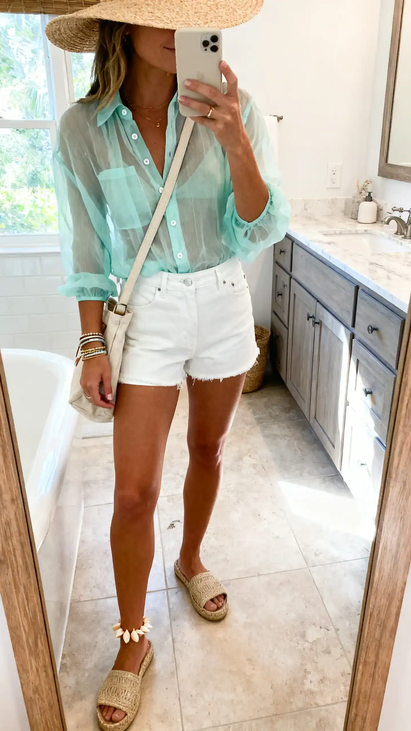 iPhone mirror selfie of a woman in a sheer seafoam gauze button-up, high-waisted white denim shorts, espadrille slides, canvas crossbody bag, shell anklet, stacked bracelets, and a wide-brim straw hat; face not visible, side angle in bathroom mirror, bright natural light, iPhone photo quality.