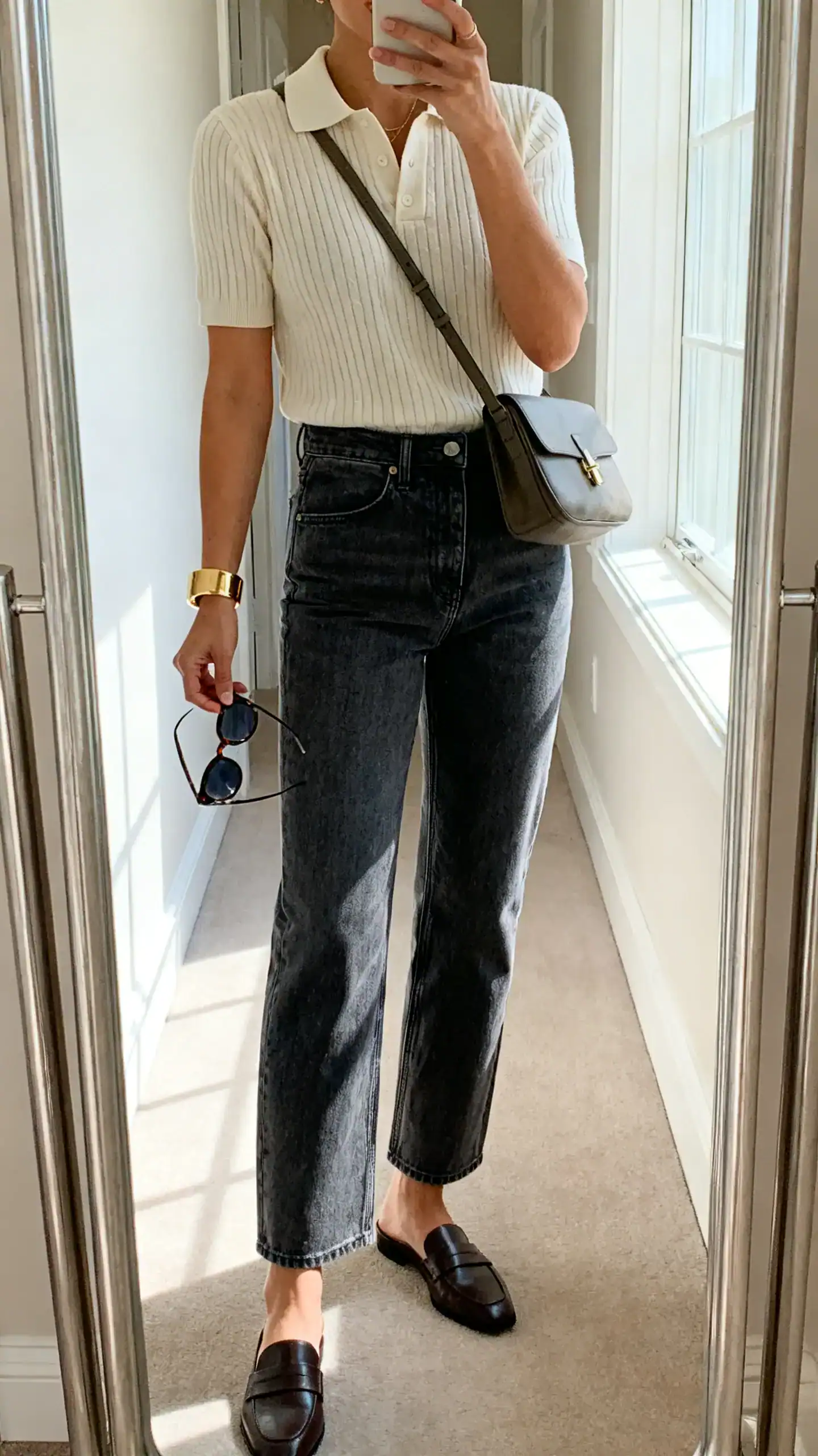 iPhone mirror selfie of a woman in a short-sleeve knit polo, high-rise straight jeans in a medium wash, loafer mules, a medium crossbody bag, gold cuff bracelet and classic sunglasses in hand, face not visible, cropped at neck, hallway full-length mirror, bright window light, iPhone photo quality.