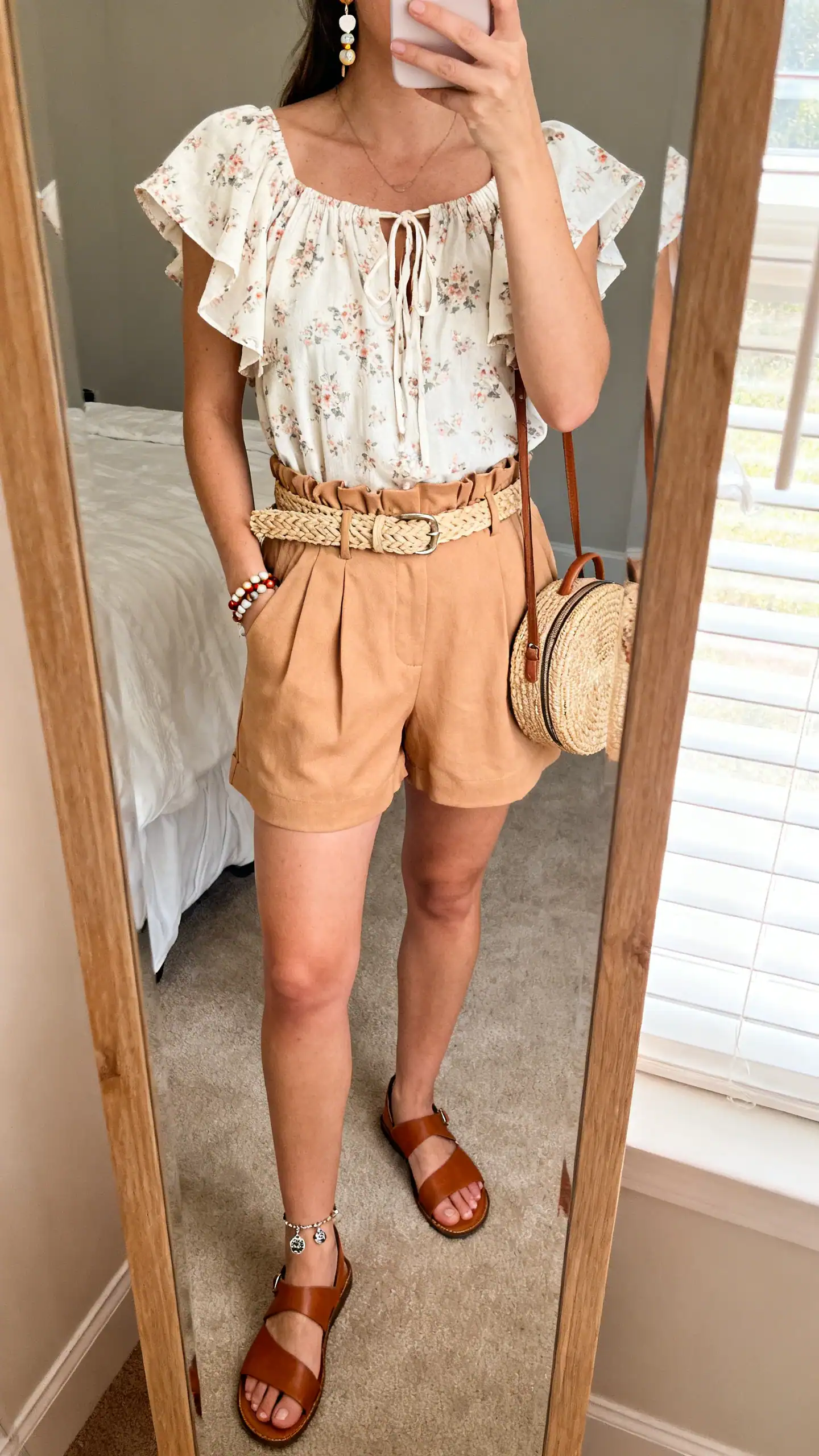 iPhone mirror selfie of a woman in a soft floral flutter-sleeve peasant top with tie neckline, high-rise tan paperbag shorts, leather toe-loop flat sandals, raffia belt, beaded earrings, charm anklet, and a compact straw backpack; face not visible, cropped at shoulders, bedroom mirror with window light, iPhone photo quality.