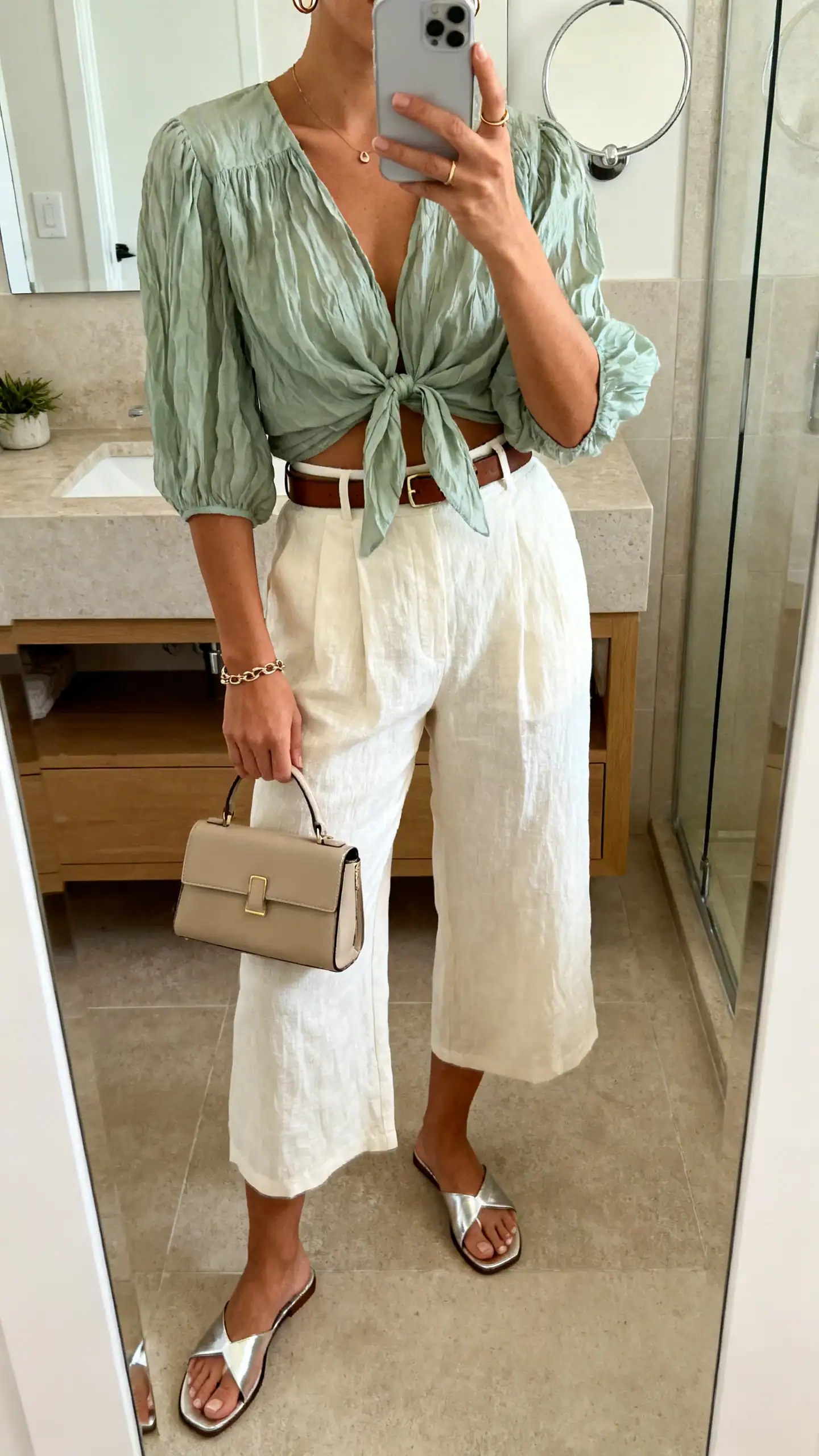 iPhone mirror selfie of a woman in a soft sage crinkled voile tie-front top, cream high-waisted linen culottes, barely-there metallic slide sandals, minimalist leather belt, tiny huggie hoops, thin chain bracelet, and a structured mini tote; face not visible, cropped at neck, bathroom mirror, soft morning light, iPhone photo quality.