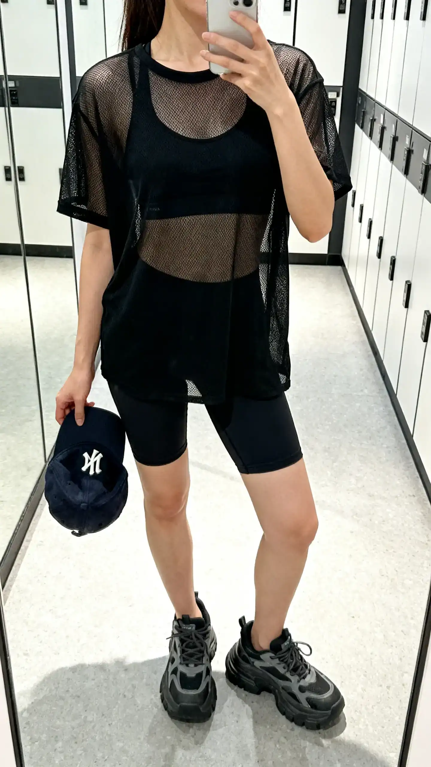 iPhone mirror selfie of a woman in a sporty-chic black mesh tee layered over a sports bra, bike shorts, chunky trainers, and a baseball cap in hand, face not visible, cropped at neck, gym locker-room mirror with bright indoor light, iPhone photo quality.