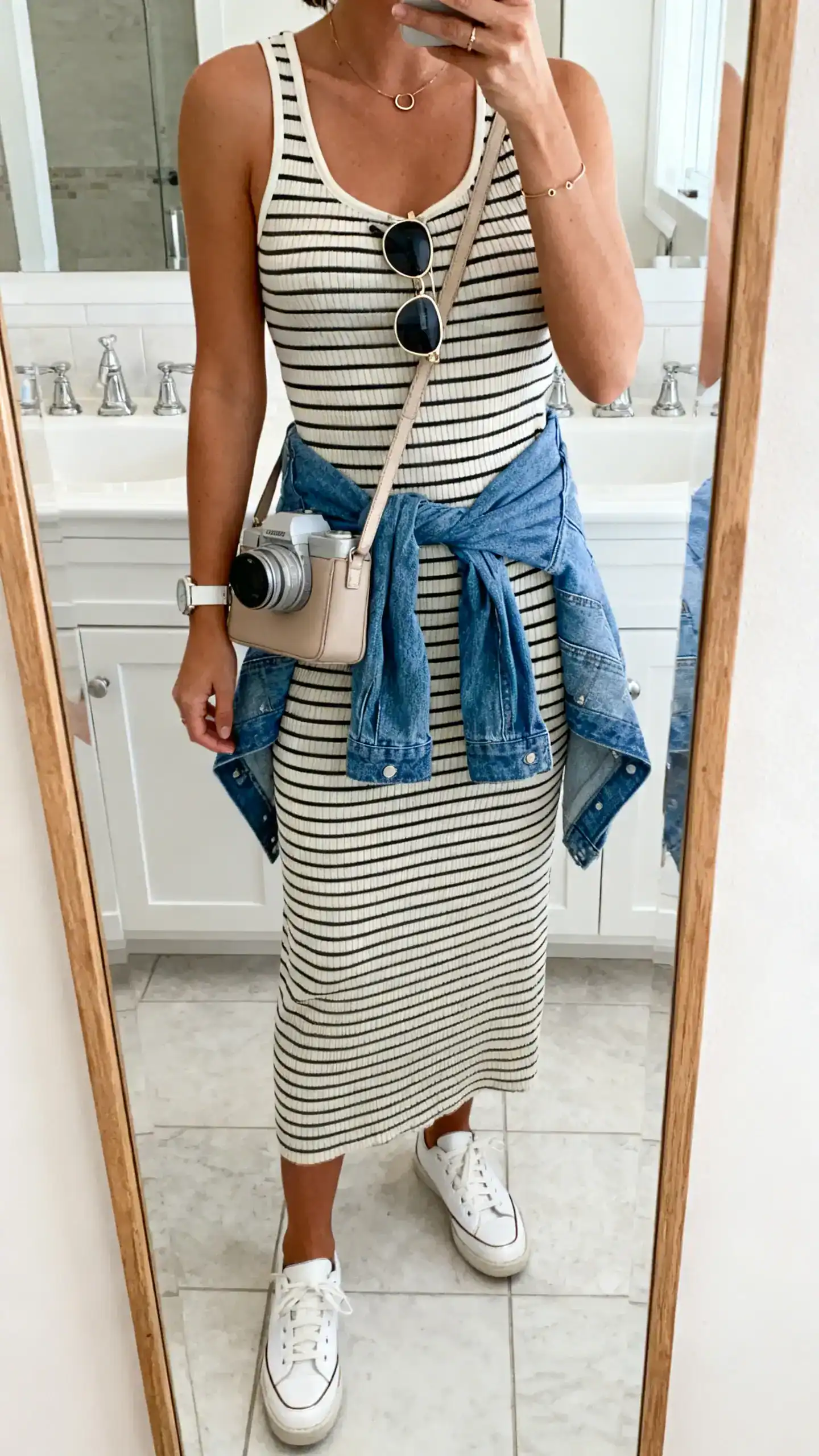 iPhone mirror selfie of a woman in a striped ribbed knit midi dress (tank style), clean white leather sneakers, a neutral camera bag crossbody, aviator sunglasses, a denim jacket tied around the waist, and a minimal watch with small hoops, face not visible, bathroom mirror, bright indoor light, iPhone photo quality.