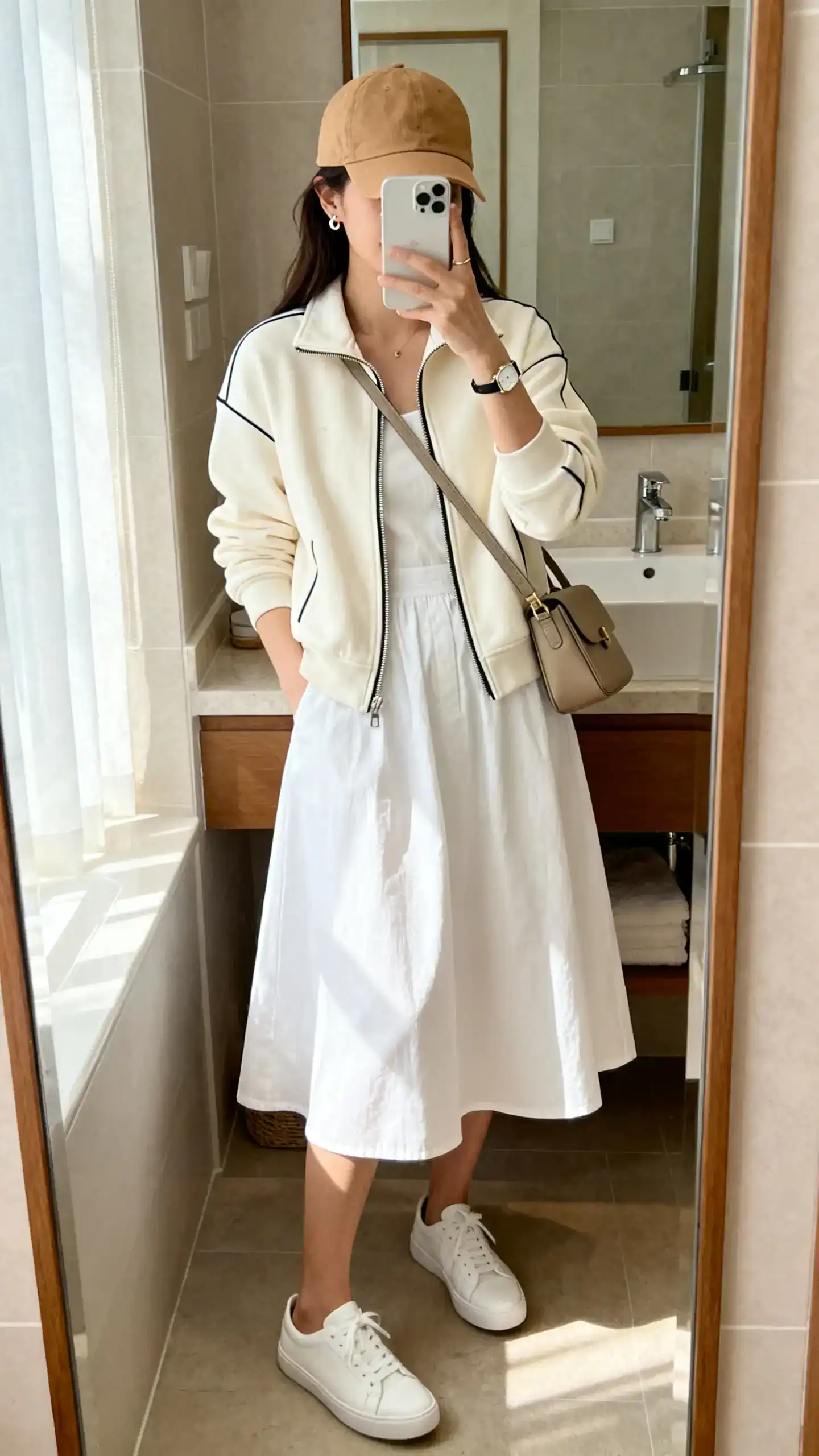 iPhone mirror selfie of a woman in a white cotton A-line sundress (knee length) with clean white low-profile sneakers, a cream zip-up track jacket, tan baseball cap, structured crossbody bag, minimal stud earrings, and a slim watch; face not visible, bathroom mirror, morning light, iPhone photo quality.