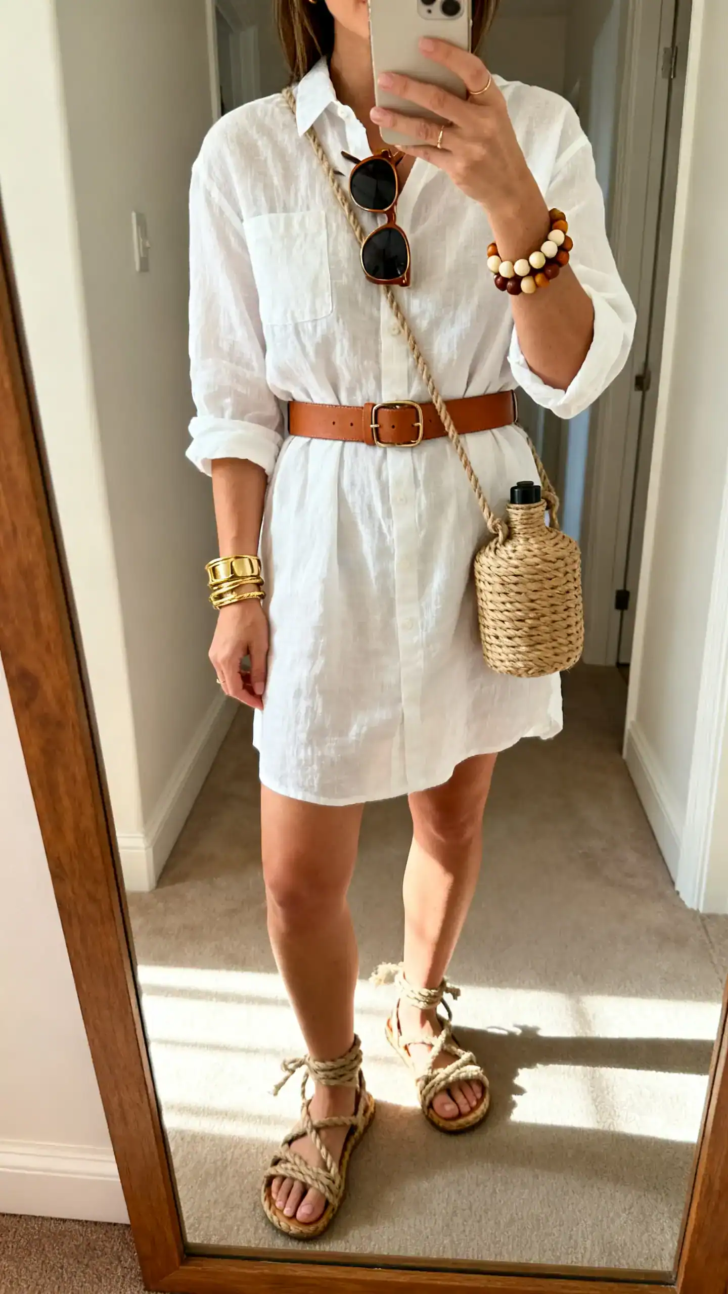 iPhone mirror selfie of a woman in a white linen shirt dress belted with a tan leather belt, rope sandals, woven canteen crossbody, oversized sunglasses, stacked gold and natural bead bracelets, face not visible, cropped at shoulders, hallway mirror with morning light, iPhone photo quality.