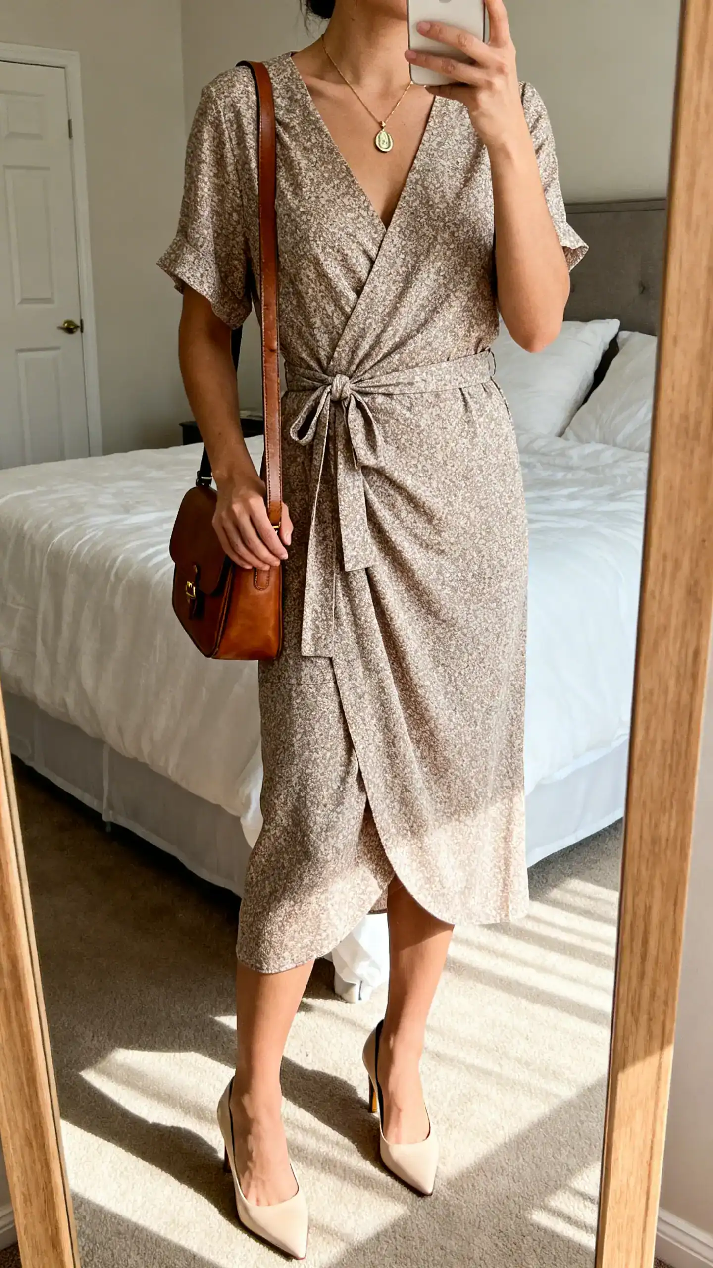 iPhone mirror selfie of a woman in a wrap dress with a V-neck and tie waist, kitten-heel pumps, pendant necklace, and a leather satchel, face not visible, cropped at neck, bedroom mirror, soft afternoon light, iPhone photo quality