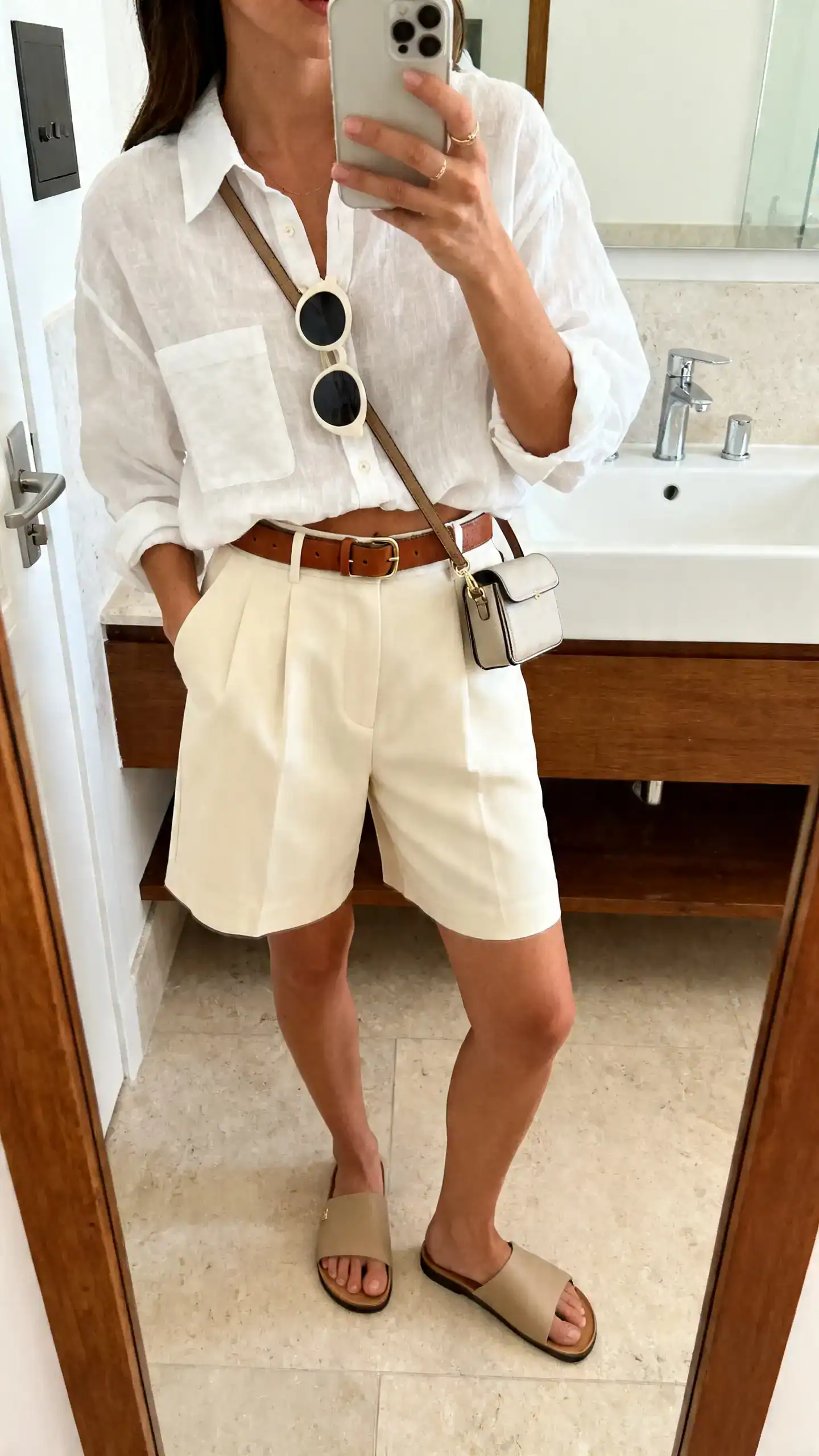 iPhone mirror selfie of a woman in high-waisted tailored shorts in cream, relaxed white linen button-down half-tucked, minimalist leather slides, a leather belt, lightweight round sunglasses on the neckline, and a compact crossbody; face not visible, cropped at shoulders, bathroom mirror, indoor light, iPhone photo quality.