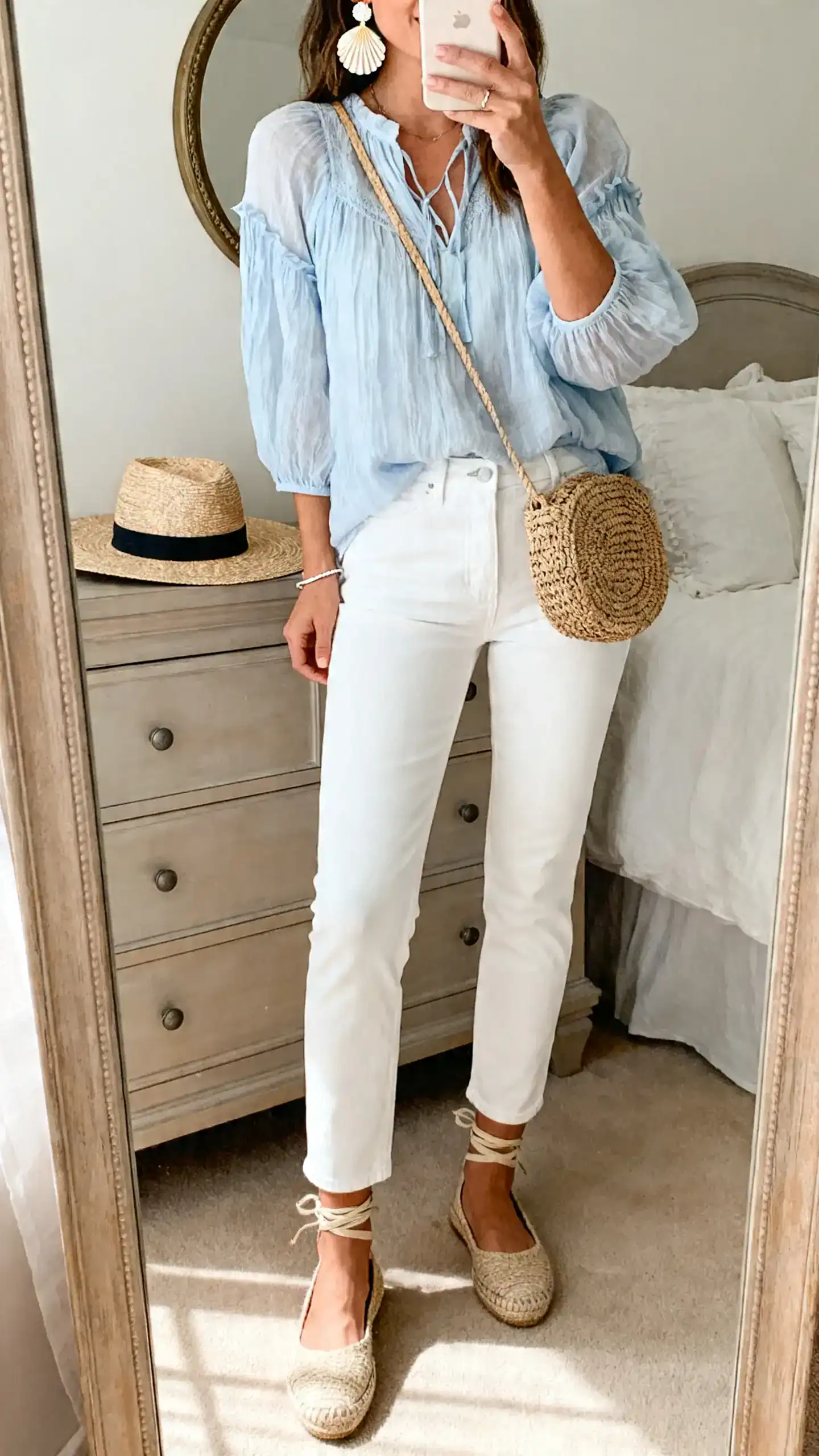 iPhone mirror selfie of a woman in straight-leg ankle-length white jeans, a sheer gauzy peasant blouse in pale blue, flat espadrilles with ankle ties, a woven crossbody, shell-inspired earrings, a delicate bracelet, and a wide-brim straw hat resting on the dresser; face not visible, cropped at shoulders, bedroom mirror, morning light, iPhone photo quality.