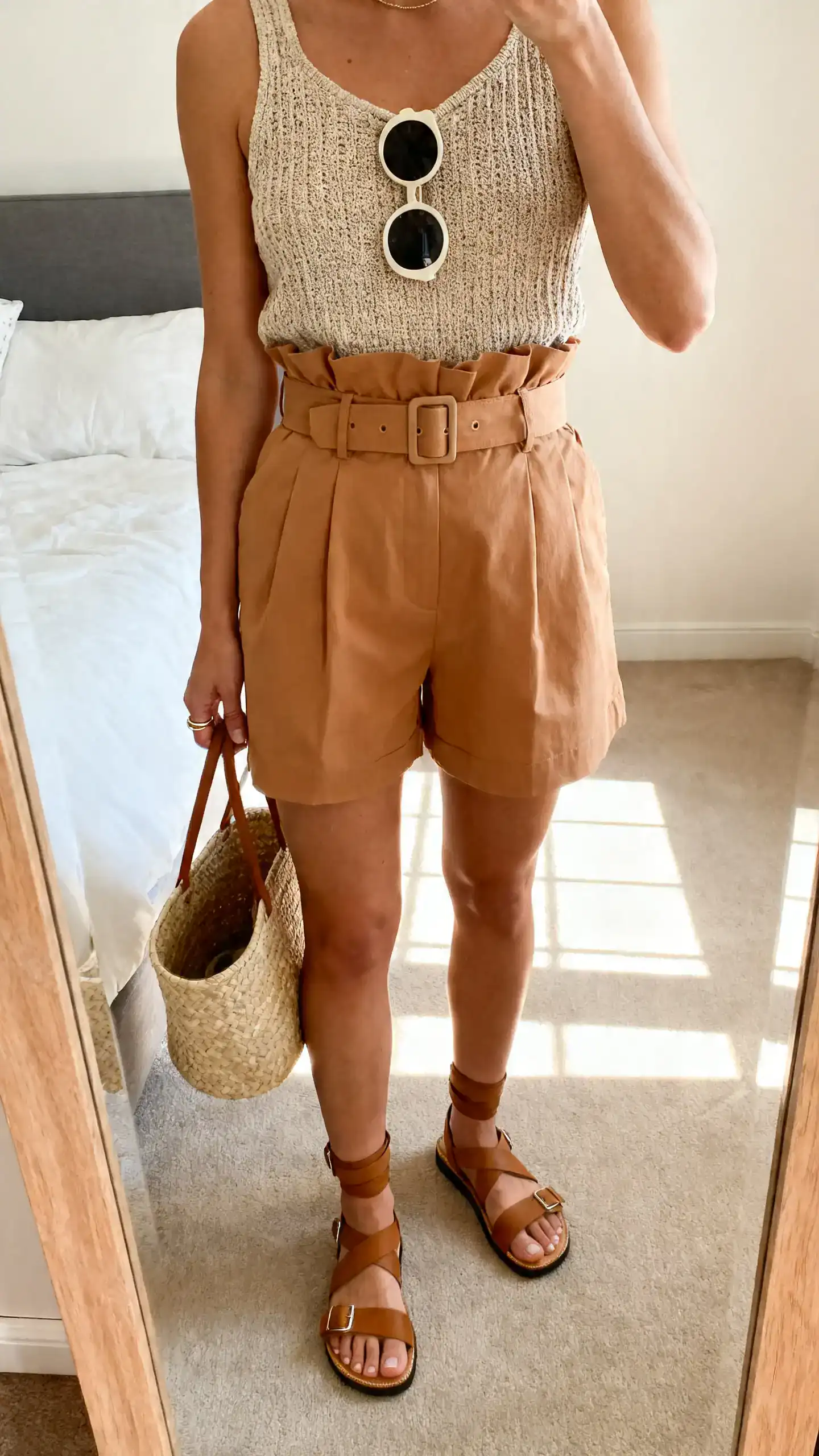 iPhone mirror selfie of a woman in tan high-waisted paperbag shorts with a compact waist belt, a textured knit tank, cushioned strappy sandals with ankle support, a raffia bucket bag, round sunglasses, and minimal stacking rings; face not visible, cropped at neck, bedroom mirror, sunny window light, iPhone photo quality.