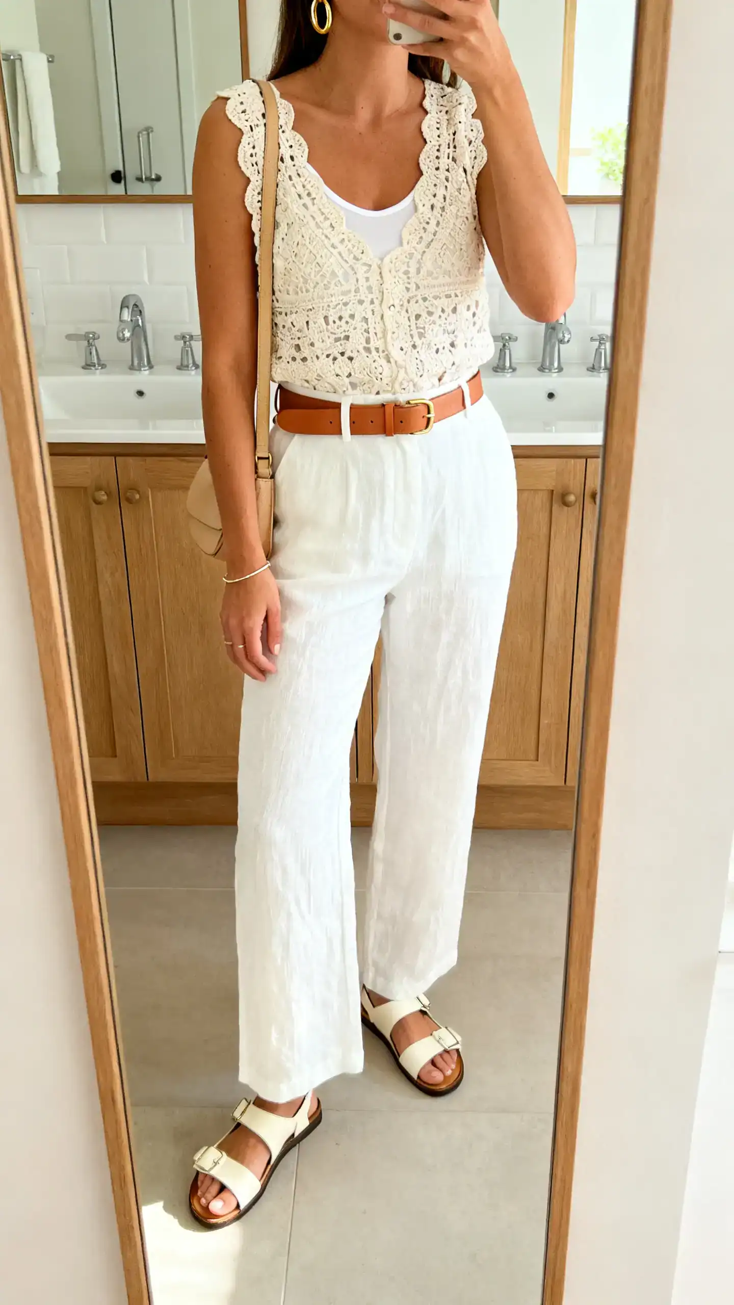 iPhone mirror selfie of a woman in white linen trousers, a lined crochet top, cream leather sandals, a tan belt, a beige crossbody, gold hoops, and a delicate bracelet, face not visible, cropped at shoulders, bathroom mirror, bright natural light, iPhone photo quality.
