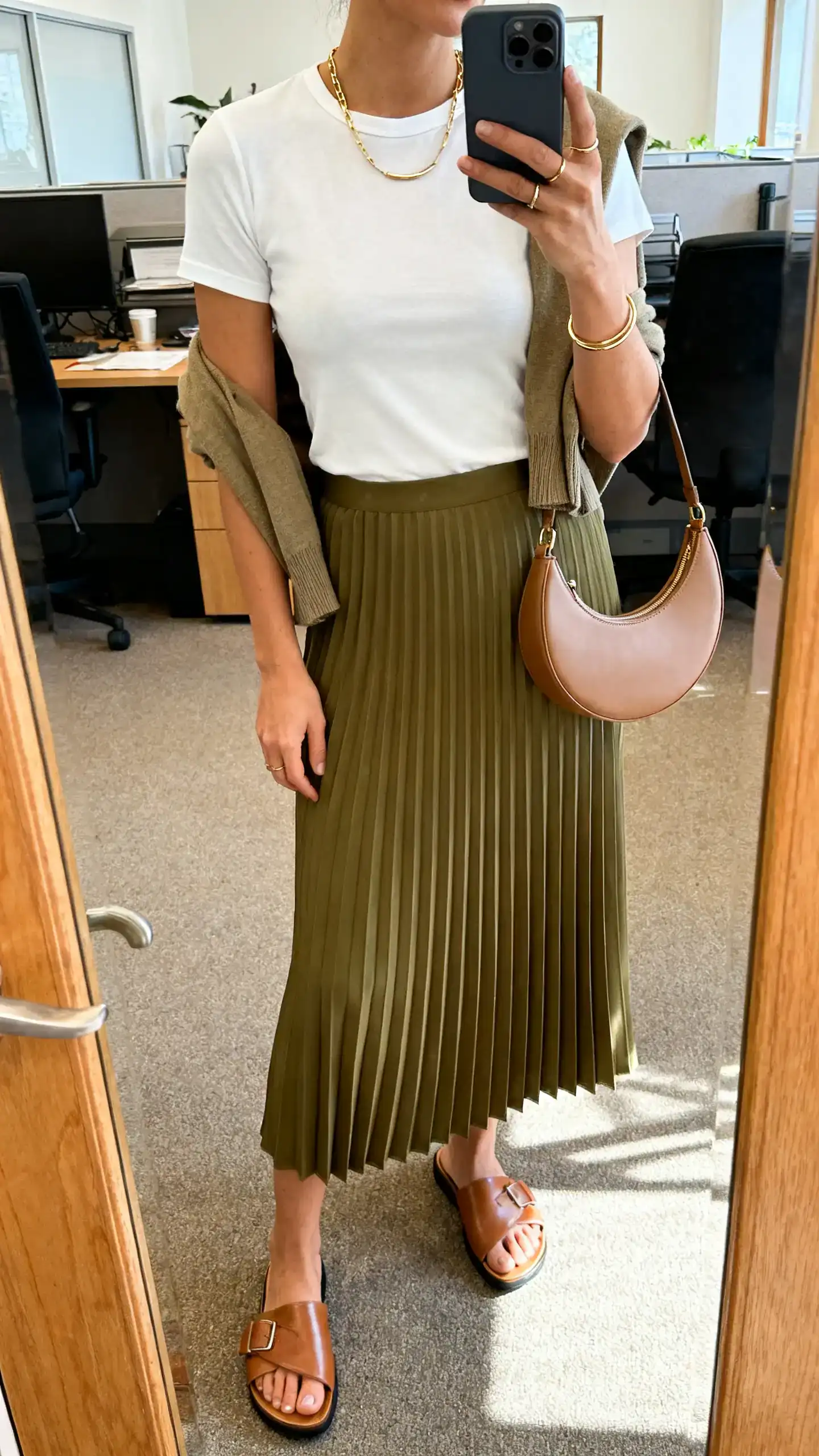 iPhone mirror selfie of a woman wearing a bias midi skirt in olive, a fitted white crewneck tee, luxe leather slides, a half-moon shoulder bag, a dainty gold chain with slim ring stack, and a cropped cardigan draped over her shoulders, face not visible, cropped at neck, office mirror, natural indoor light, iPhone photo quality.