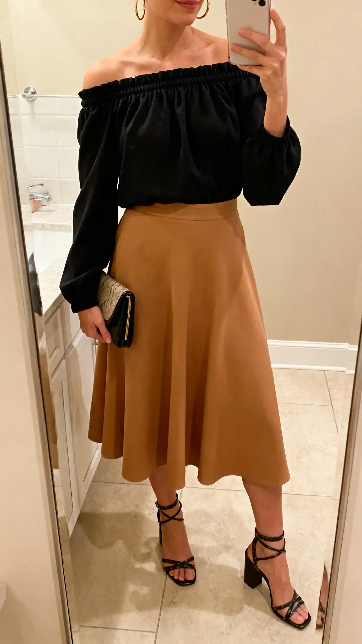 iPhone mirror selfie of a woman wearing a black off-shoulder top with elastic neckline, A-line midi skirt in tan, strappy block-heel sandals, hoop earrings, clutch, face not visible, cropped at neck, bathroom mirror, warm indoor lighting, iPhone photo quality