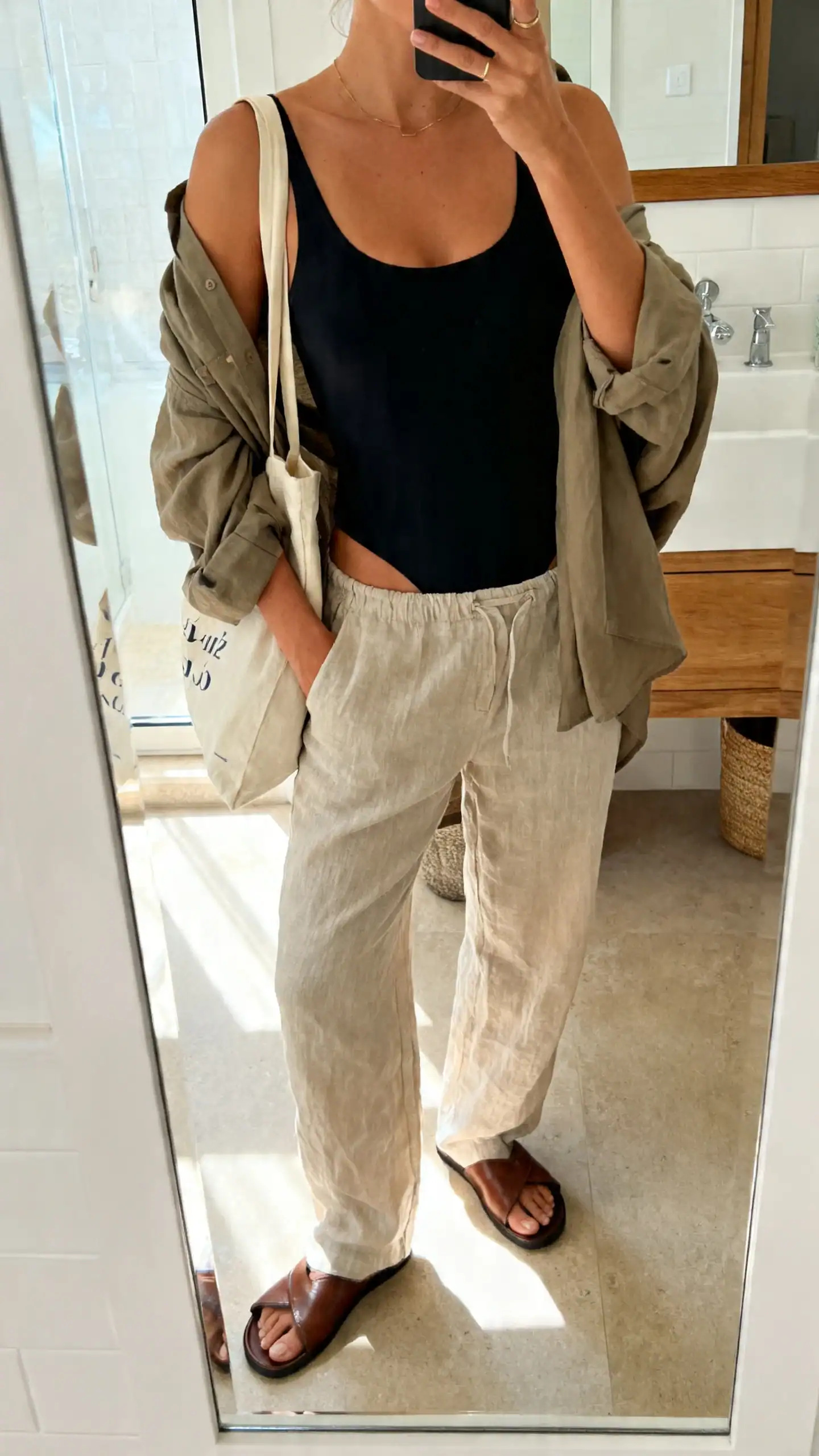 iPhone mirror selfie of a woman wearing a black one-piece swimsuit with relaxed linen trousers, leather slides, oversized shirt draped over shoulders, tote bag, face not visible, cropped at neck, bathroom mirror, bright natural light, iPhone photo quality
