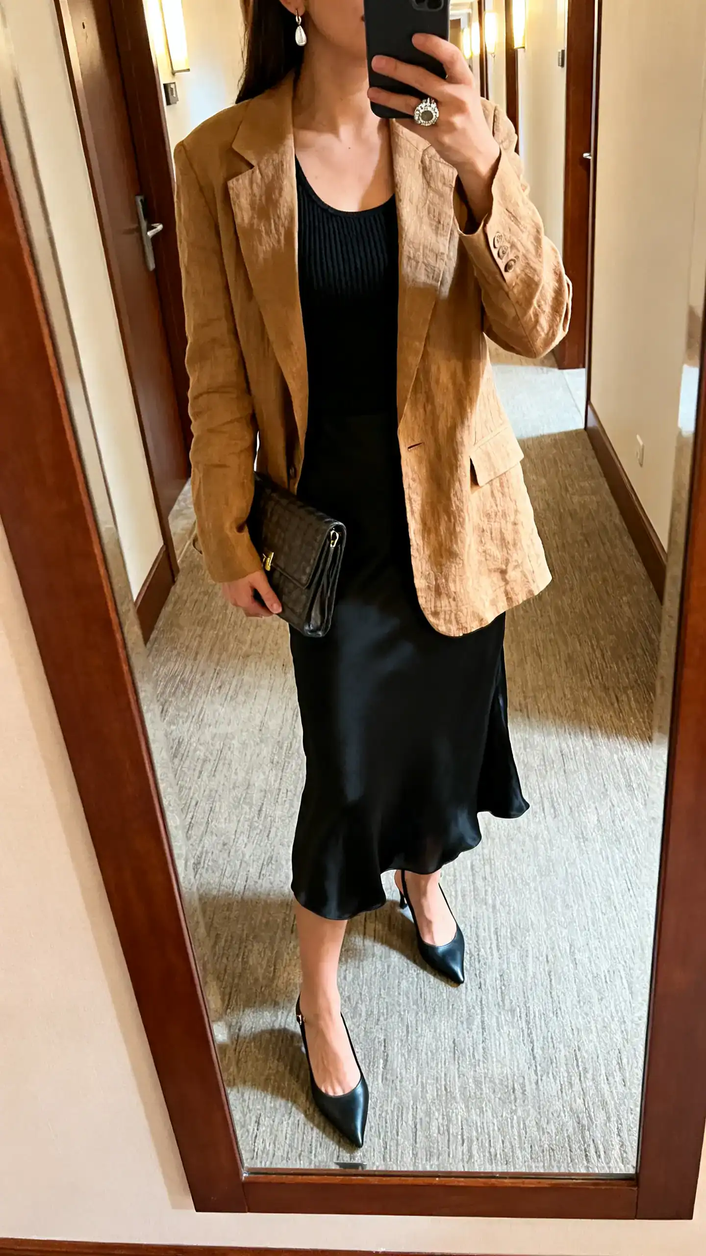 iPhone mirror selfie of a woman wearing a black slip midi skirt, a fitted knit tank, a camel linen blazer, slingback kitten heels, a structured clutch, a statement ring and subtle drop earrings, face not visible, cropped at neck, hotel hallway mirror, warm indoor lighting, iPhone photo quality.