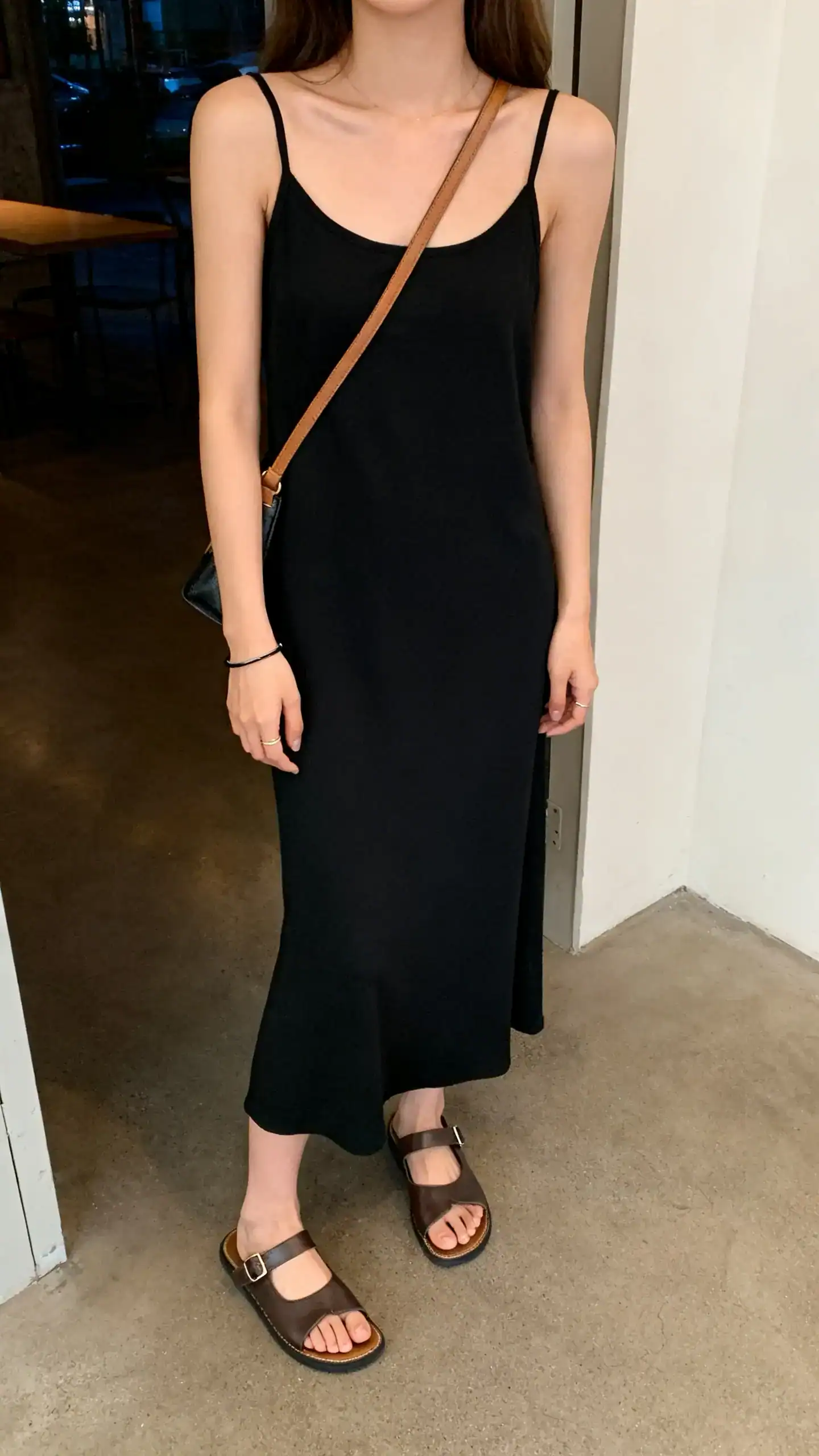 iPhone mirror selfie of a woman wearing a black slip dress with flat leather sandals, crossbody bag, and simple bracelet, face not visible, cropped at neck, evening indoor light, iPhone photo quality