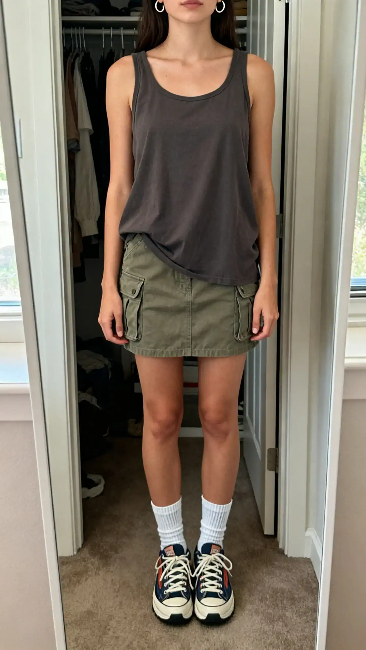 iPhone mirror selfie of a woman wearing a boxy tank and a cargo mini skirt with crew socks, retro sneakers, and small hoop earrings, face not visible, cropped at neck, closet mirror, natural daylight, iPhone photo quality.