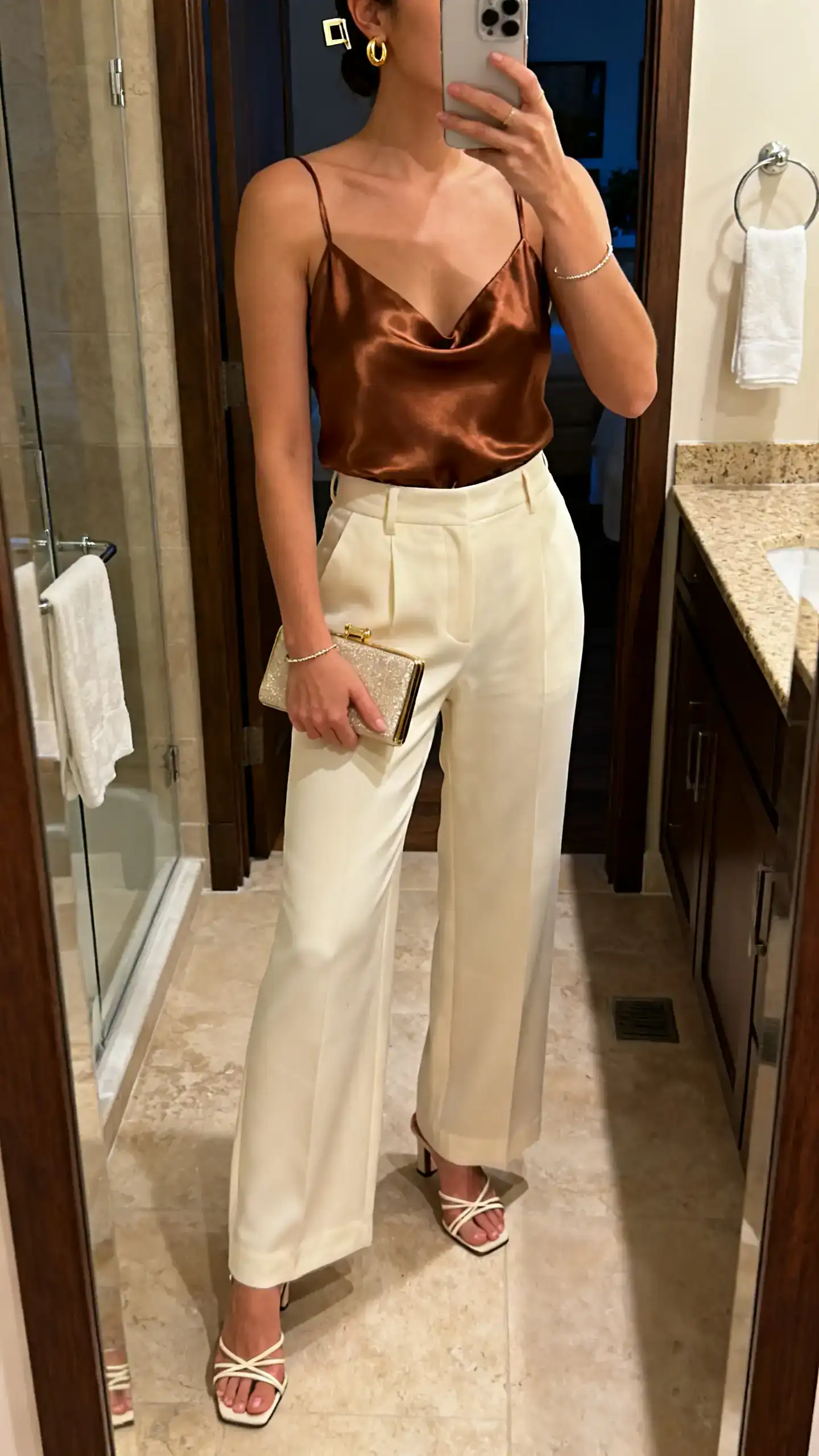 iPhone mirror selfie of a woman wearing a bronze satin camisole with high-waisted cream wide-leg trousers, strappy heeled sandals, compact clutch, gold huggies, delicate bracelet, sleek hair clip, face not visible, cropped at neck, bathroom mirror, warm indoor evening light, iPhone photo quality.