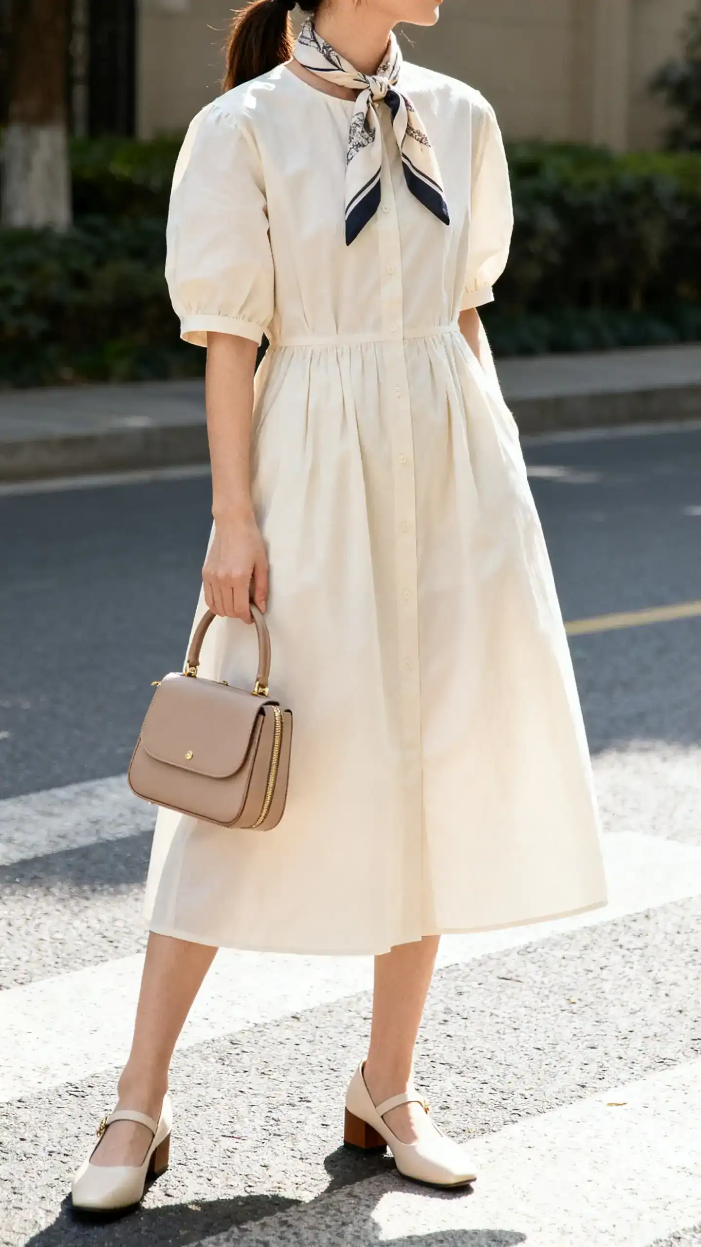 iPhone mirror selfie of a woman wearing a cotton poplin dress with low block heels, scarf-tied pony, and a compact top-handle bag, face not visible, cropped at shoulders, bright morning light, iPhone photo quality