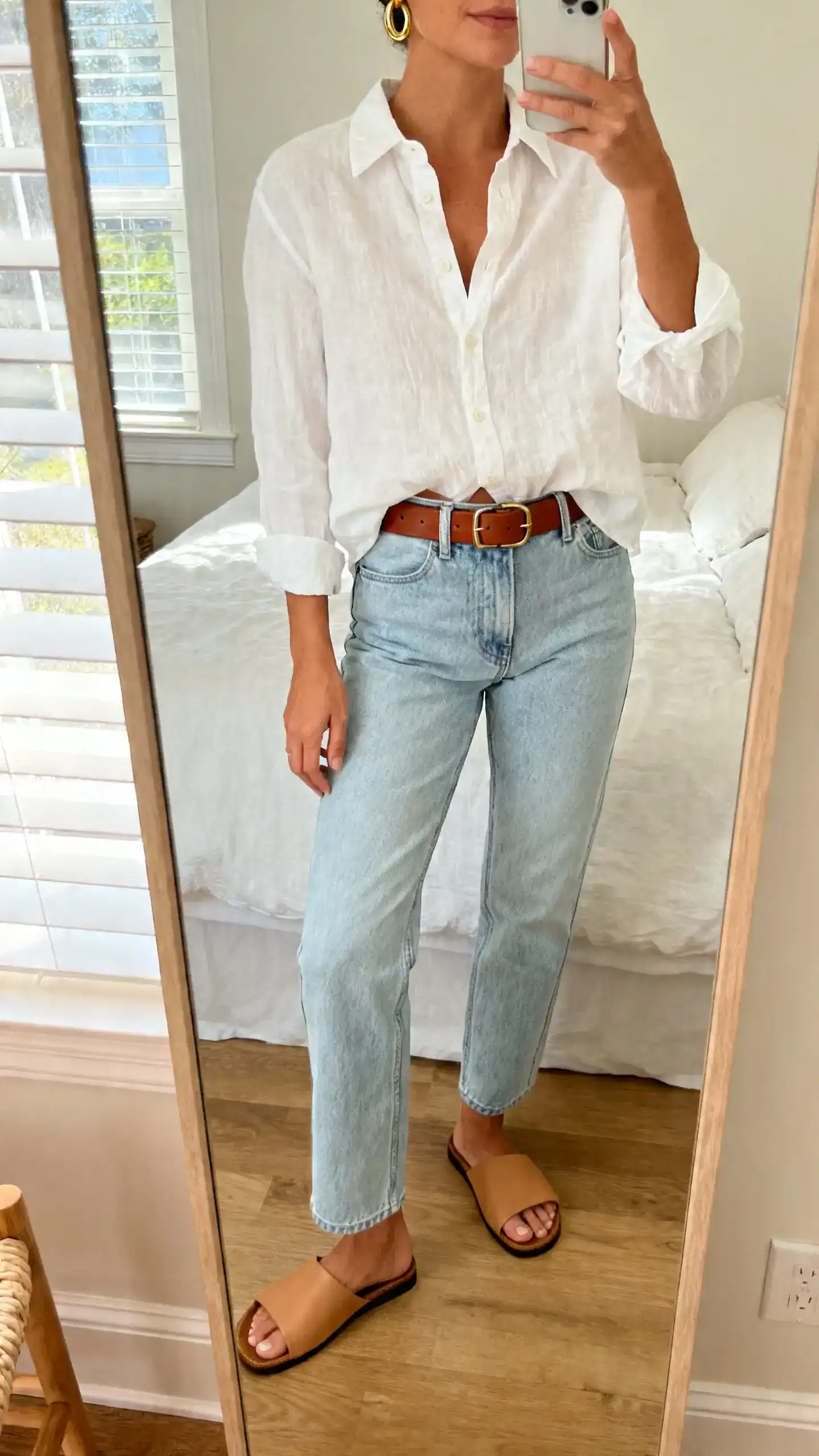 iPhone mirror selfie of a woman wearing a crisp white linen button-up shirt half-tucked into light wash straight-leg jeans with brown leather belt, simple gold hoops, tan slide sandals, face not visible, cropped at neck, bedroom mirror, morning window light, iPhone photo quality