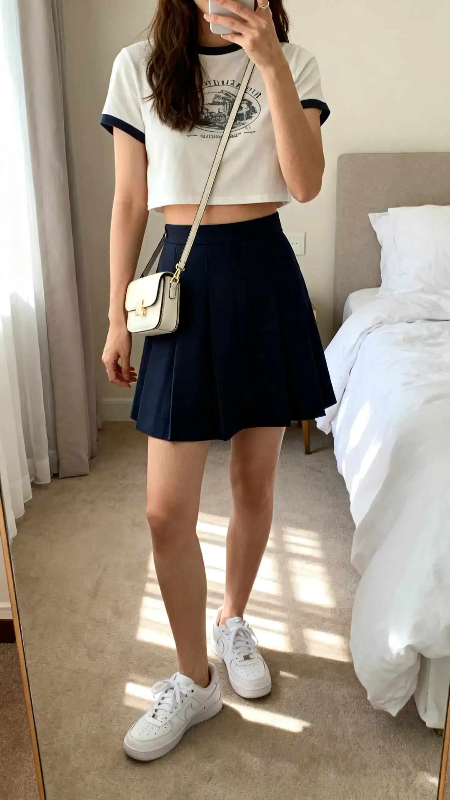 iPhone mirror selfie of a woman wearing a cropped tee with a high-waisted A-line skirt, white sneakers, and a mini crossbody for gelato runs, face not visible, cropped at neck, casual bedroom setting, iPhone photo quality